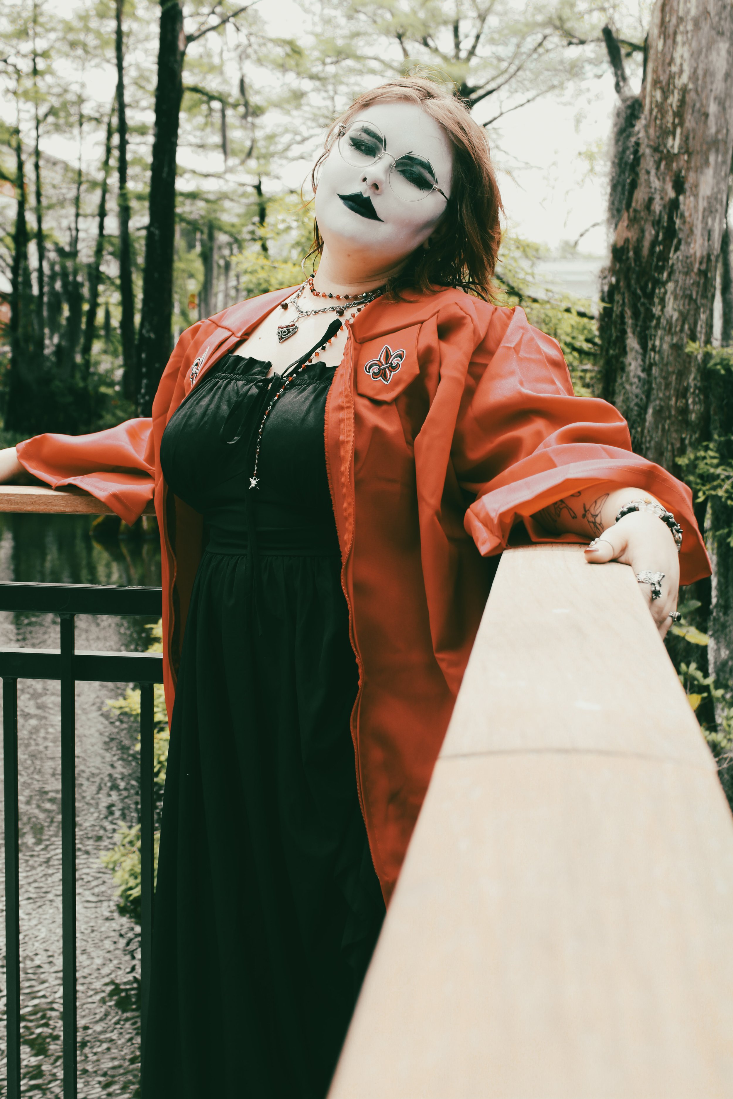A woman with face makeup dressed in black and red clothing, wearing sunglasses, jewelry, and makeup, standing on a wooden bridge in a wooded area.