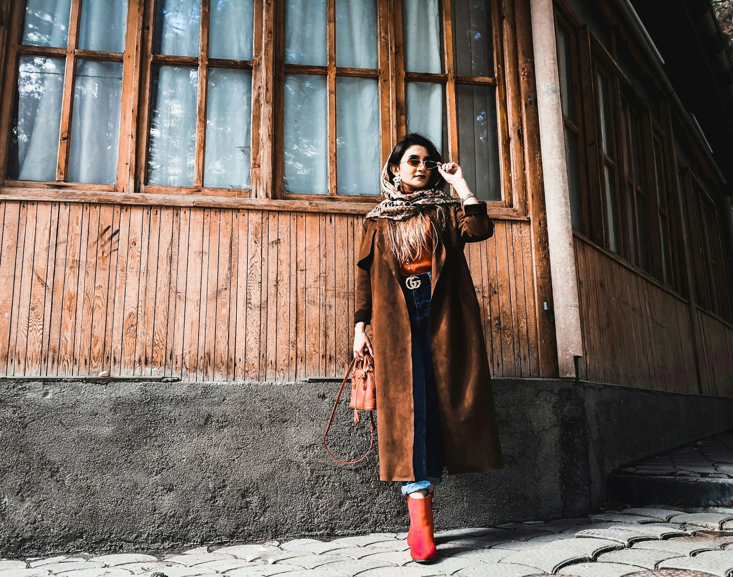Woman standing in front of a wooden wall holding sunglasses and wearing a brown coat, scarf, jeans, and red boots.