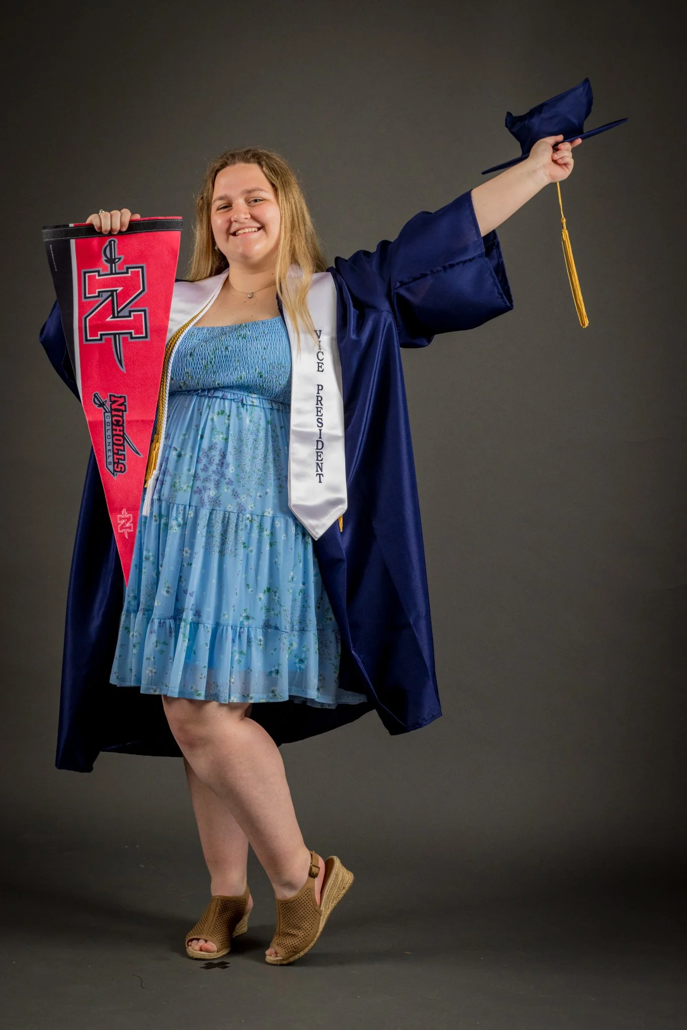 A young woman in a blue floral dress and tan wedge shoes holding up a graduation cap and a university pennant, smiling at the camera.