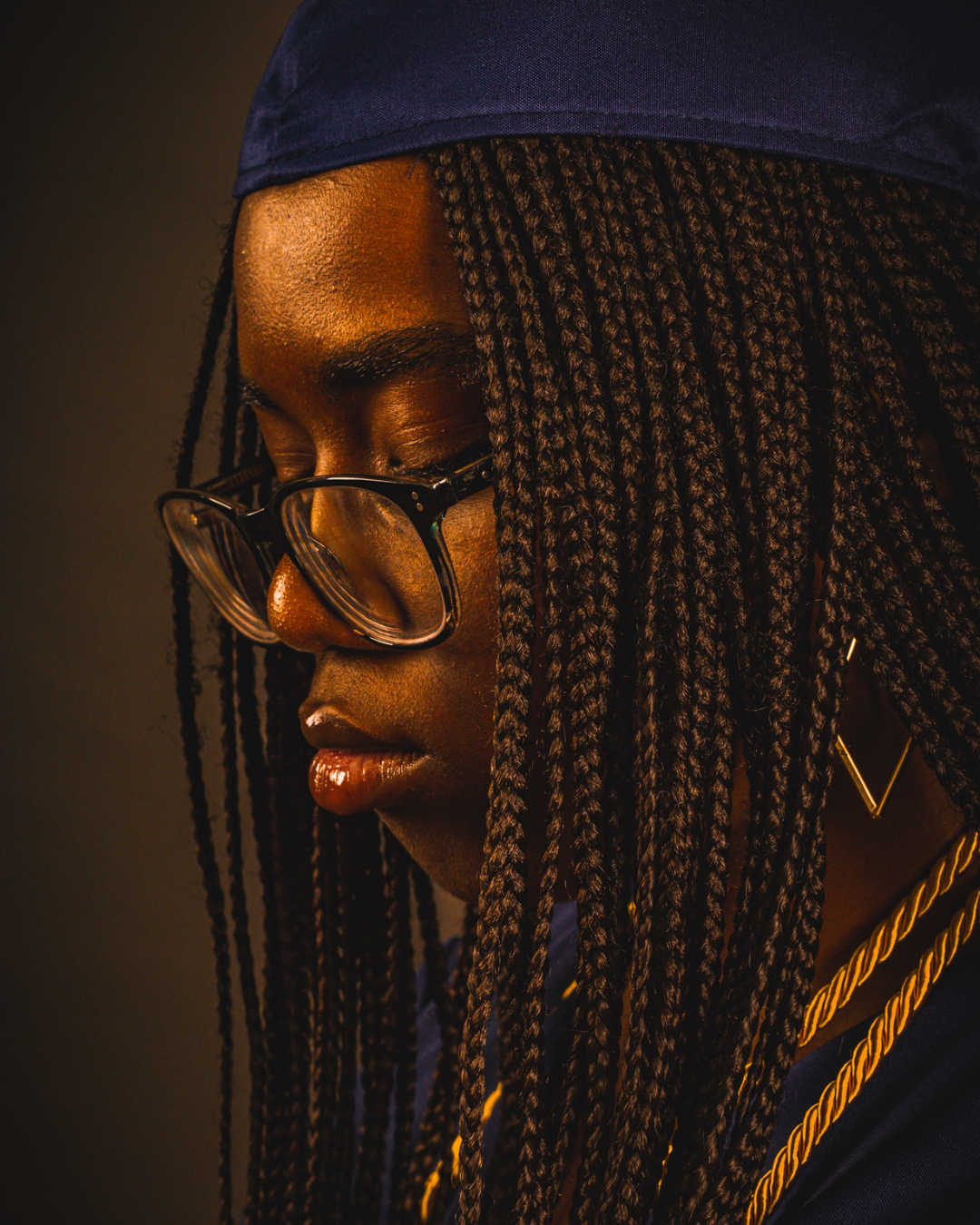 A young woman with dark skin and long braided hair wearing glasses and a navy graduation cap and gown, looking downward.