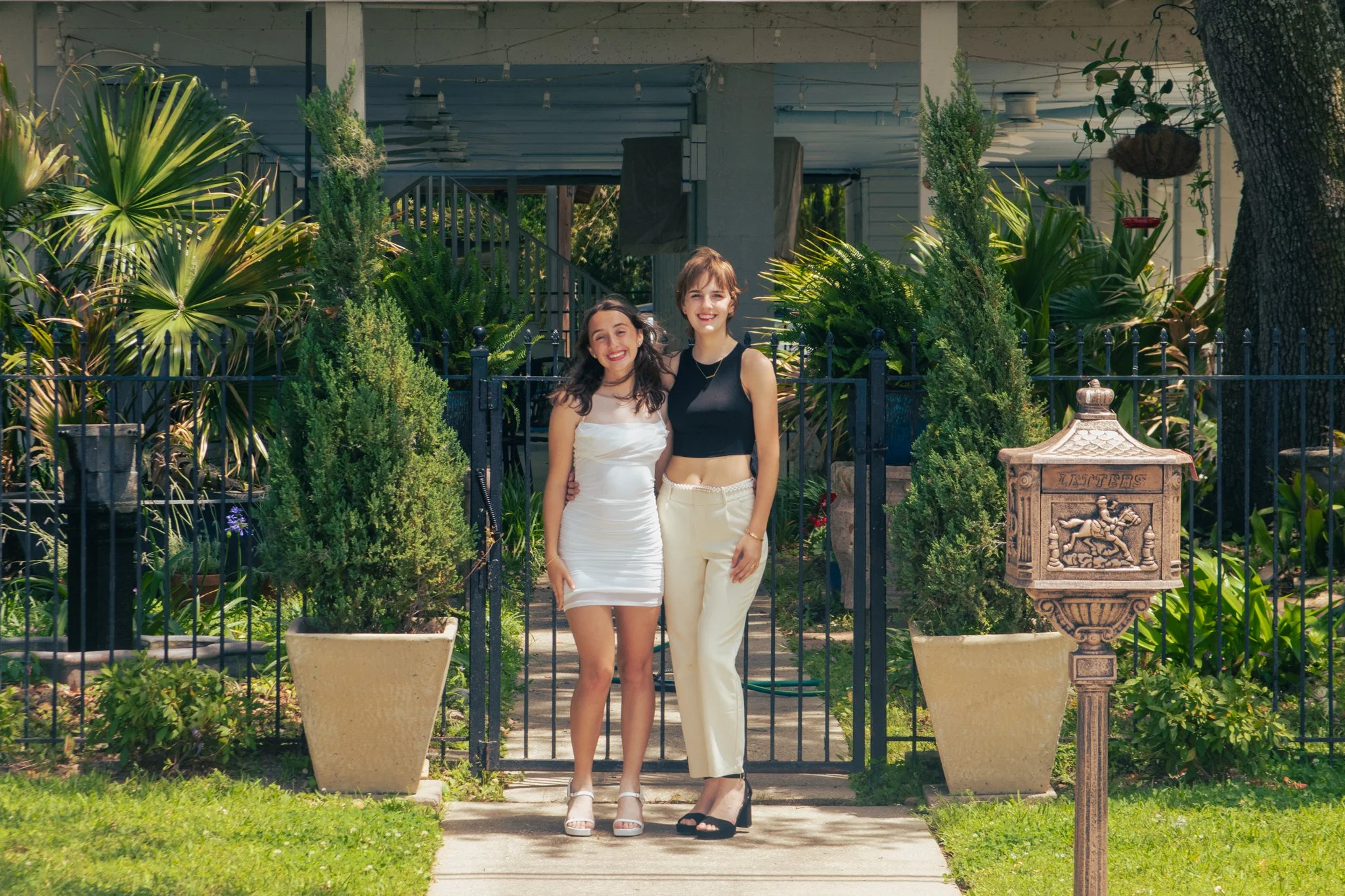 Two young women standing on a sidewalk in front of a house with an iron fence and lush greenery. They are smiling and posing together, one in a white dress and the other in a black top and beige pants.