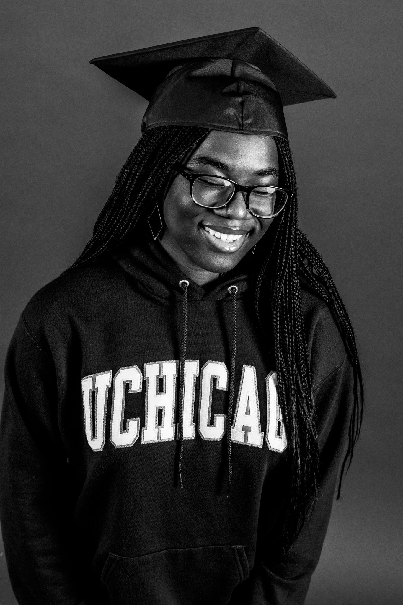 A woman wearing a graduation cap and glasses, smiling, dressed in a USC Chicago hoodie, in a studio portrait.