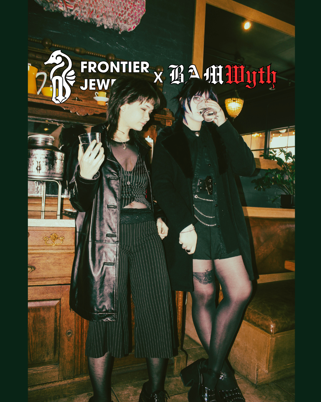 Two women in dark, edgy fashion, standing in a cozy, vintage-style café with wooden furniture and warm lighting. One woman on the left is wearing a black leather coat, striped pants, and holding a glass, while the woman on the right is wearing a blac