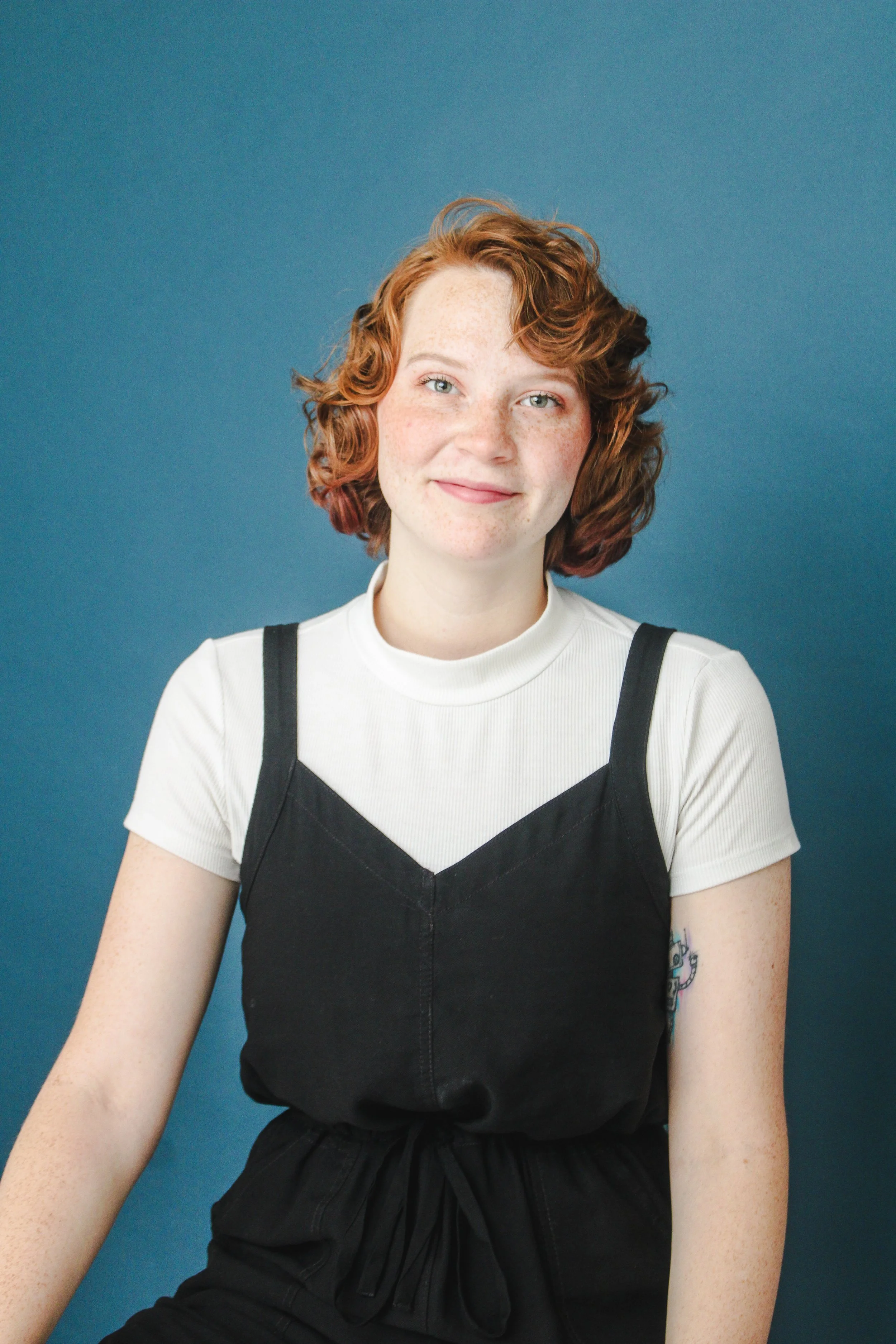 Portrait of a young woman with curly red hair, freckles, and blue eyes, wearing a white t-shirt and black overalls, sitting against a solid blue background.