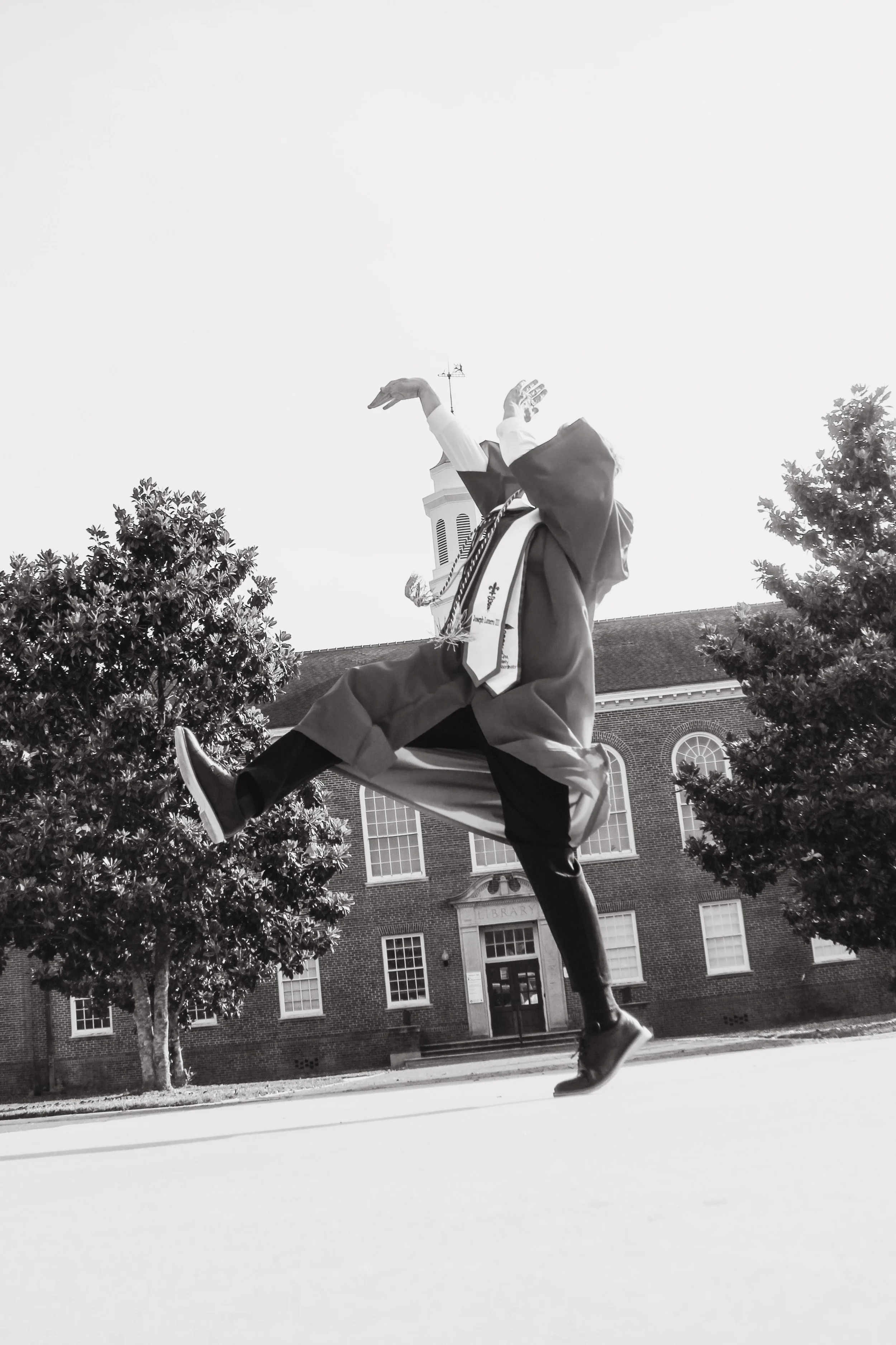 A black-and-white photo of a person on a scooter in front of a brick building with a steeple. The person is wearing a graduation gown and lei, with an arm raised above the head and the other bent at the elbow. Trees frame the scene.