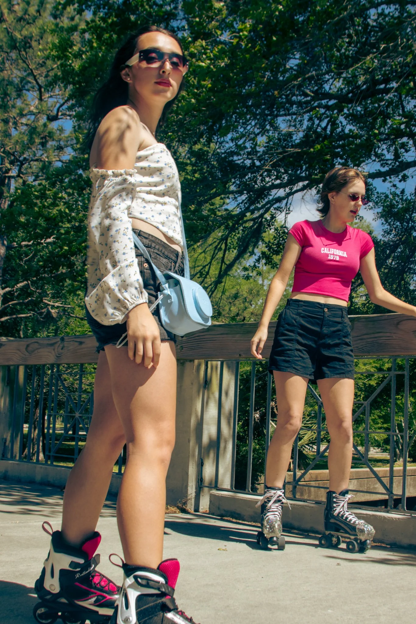 Two young women roller skating outdoors on a sunny day. One in the foreground with sunglasses, a floral off-shoulder top, and black shorts, holding a small light blue bag. The other in the background wearing a pink crop top, black shorts, and sunglas