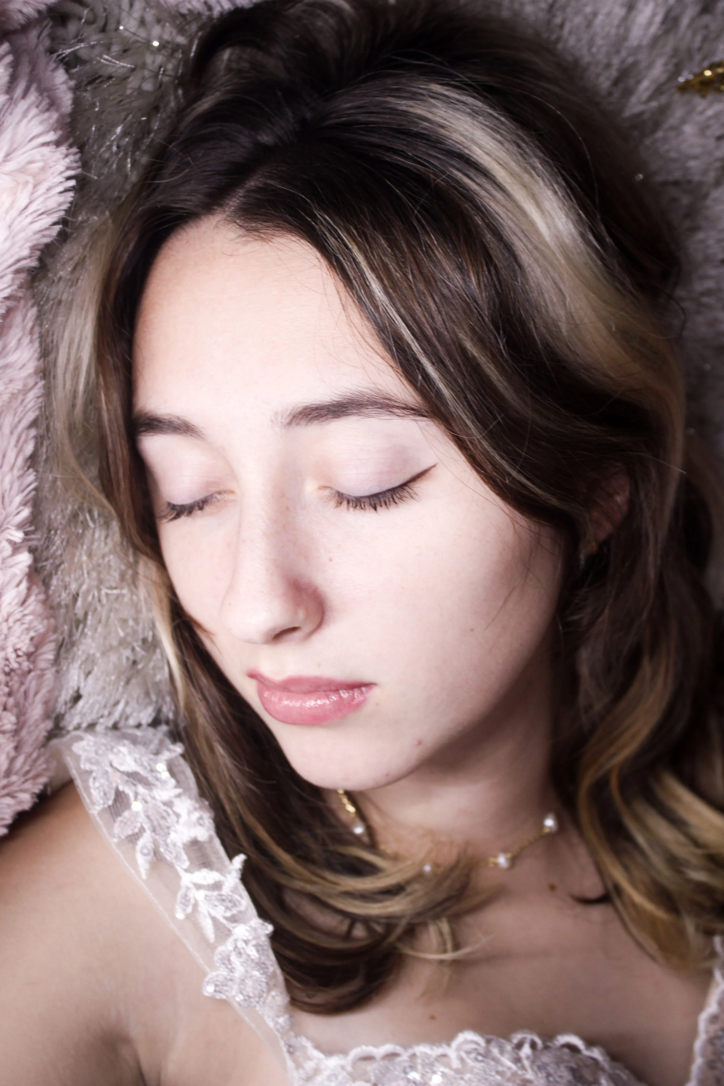 Close-up of a young woman with brown hair and light makeup, lying on her side with her eyes closed, surrounded by soft blankets.