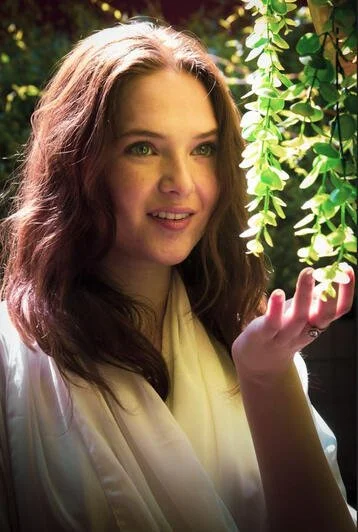 A young woman with long, wavy brown hair and green eyes standing outdoors near green leafy plants, looking at the leaves while holding one in her hand.