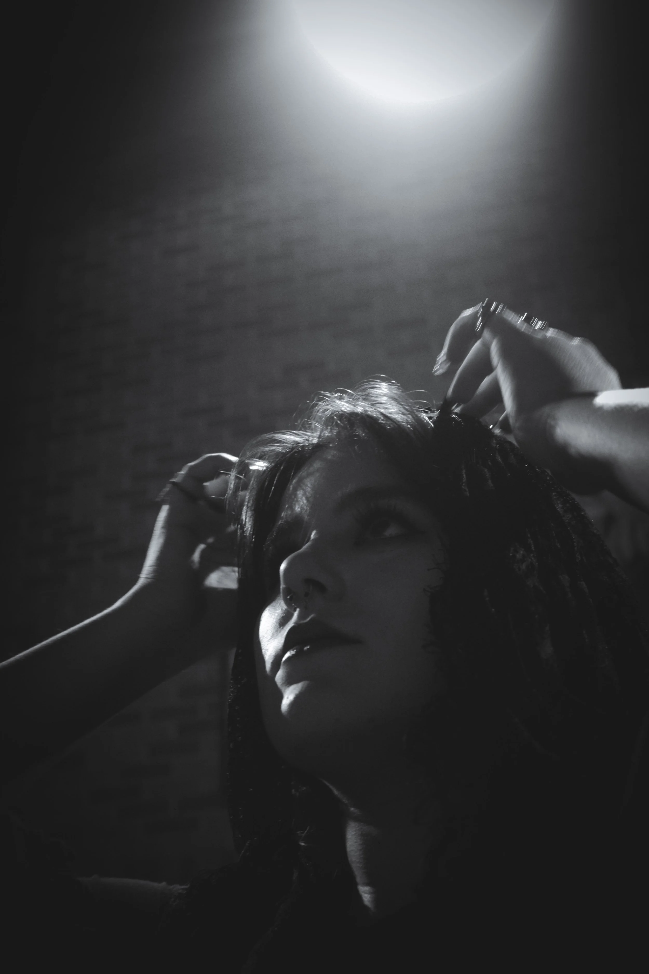 A black-and-white photograph of a woman looking upward, with her hands in her hair, illuminated by a bright overhead light against a dark background.