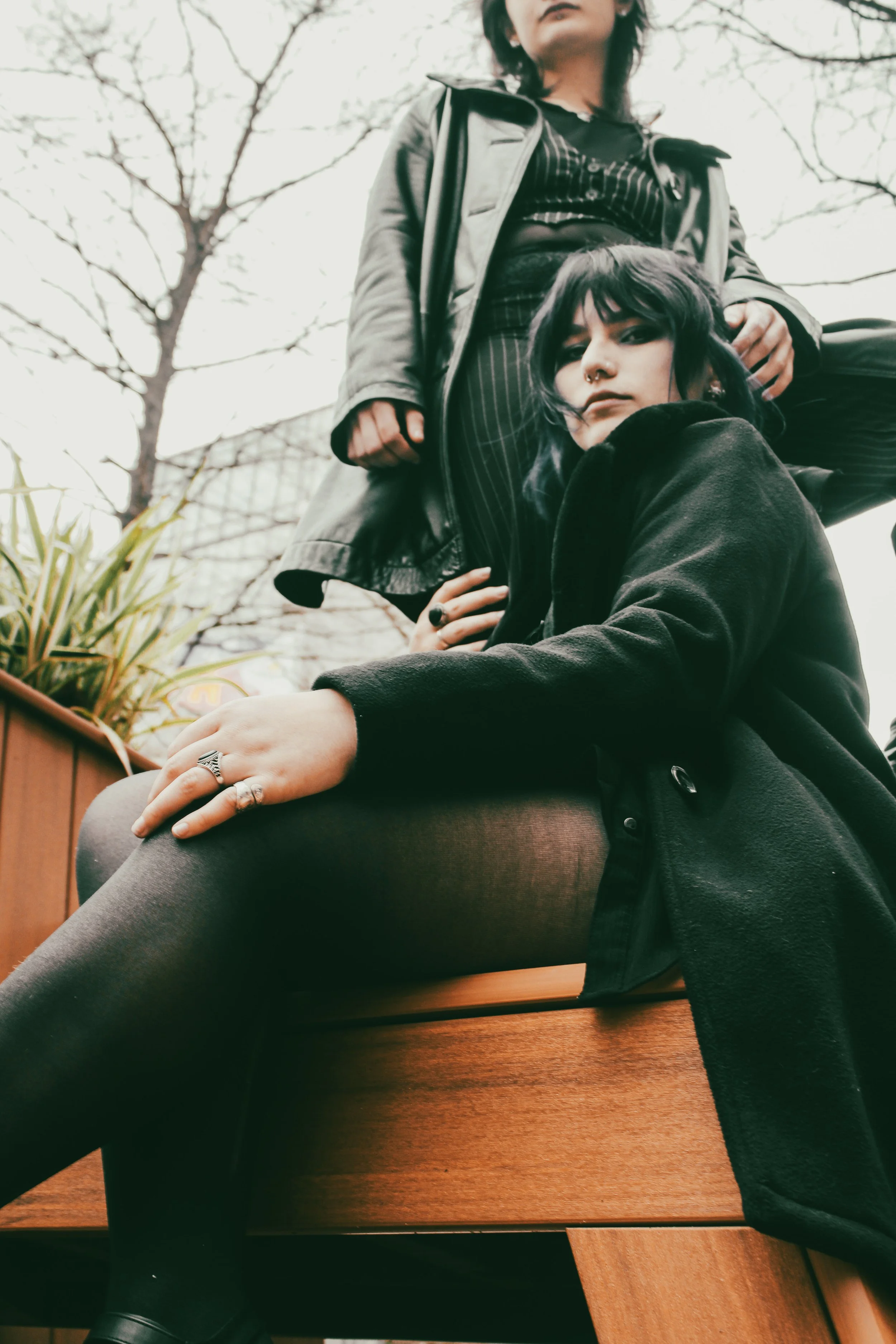 Two women with dark hair and punk style clothing outdoors on a cloudy day, one sitting on a wooden bench and the other standing behind her, with leafless trees in the background.