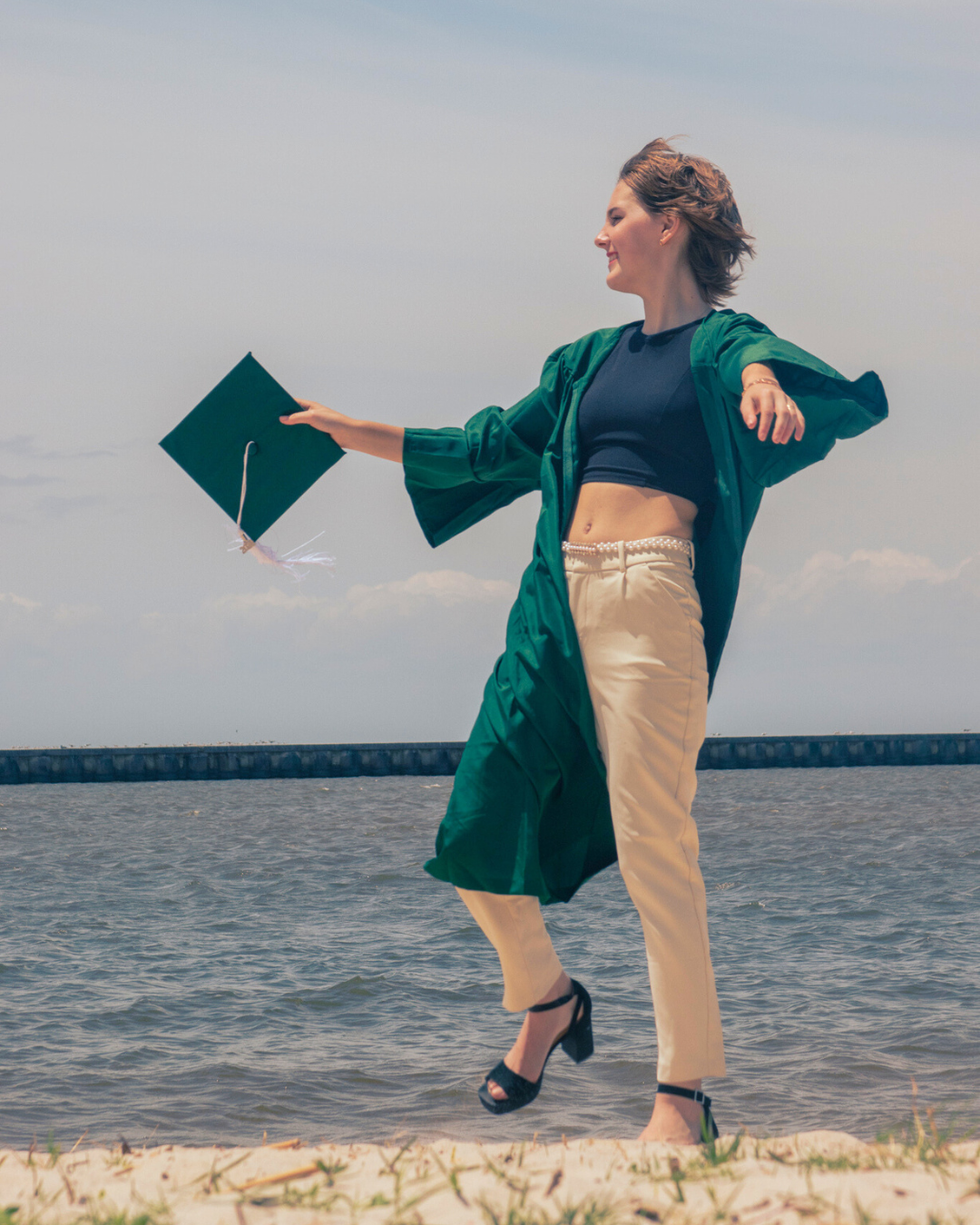 A young woman in a graduation gown joyfully throwing her cap while standing on a beach with water and a cloudy sky in the background.