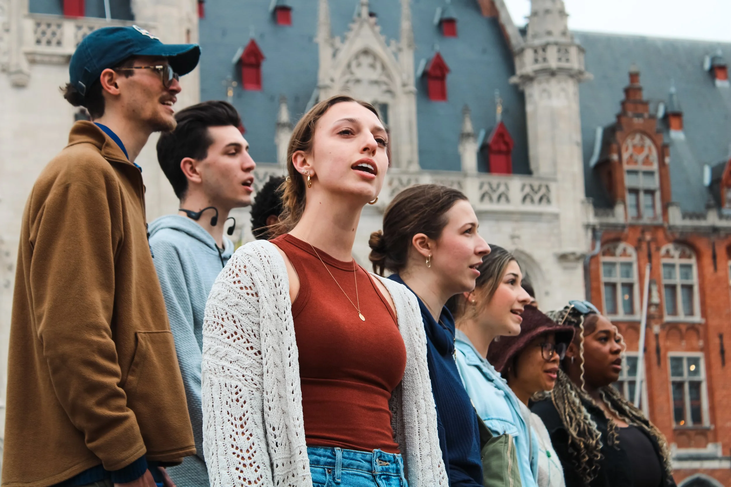 A diverse group of people standing outdoors in front of a historic building, possibly singing or listening attentively.