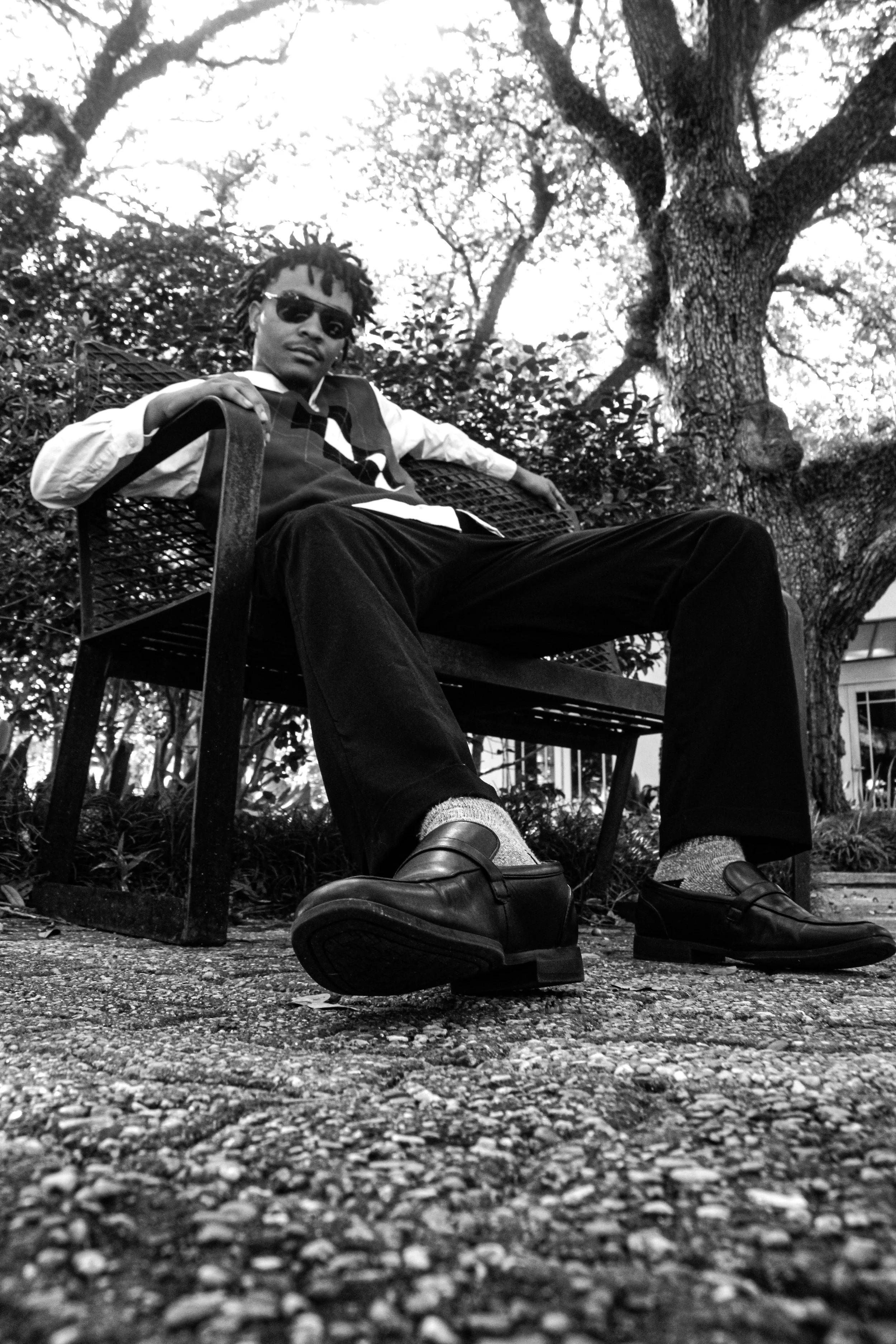 A man with dreadlocks wearing sunglasses, a patterned vest, dress shirt, and dress shoes sitting on a metal bench outdoors underneath a large tree.