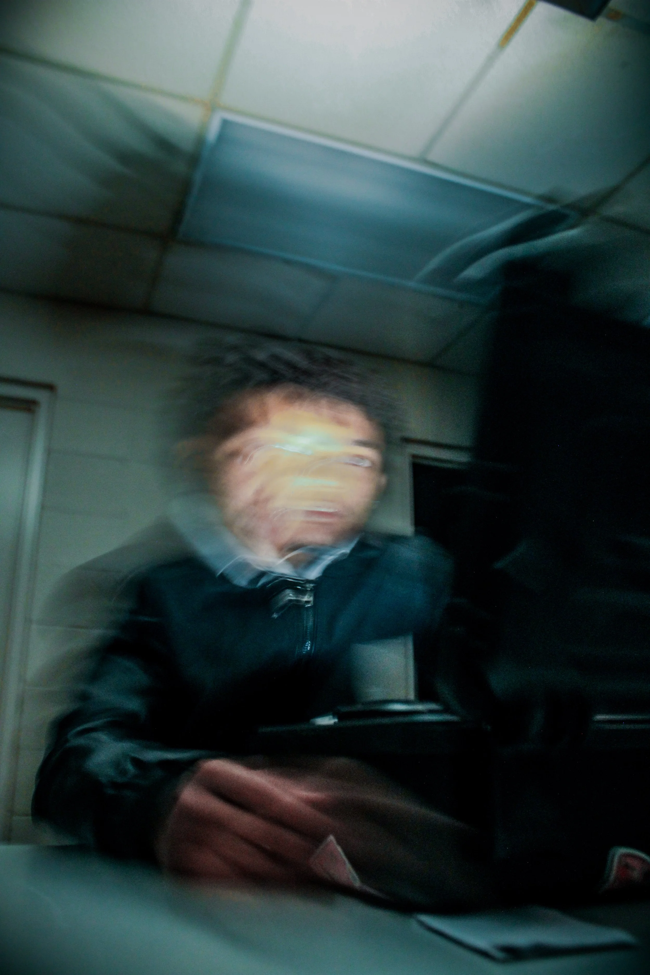 Blurred image of a person wearing glasses and a dark jacket, sitting in a room with a tiled ceiling, holding an object, with a dark background.