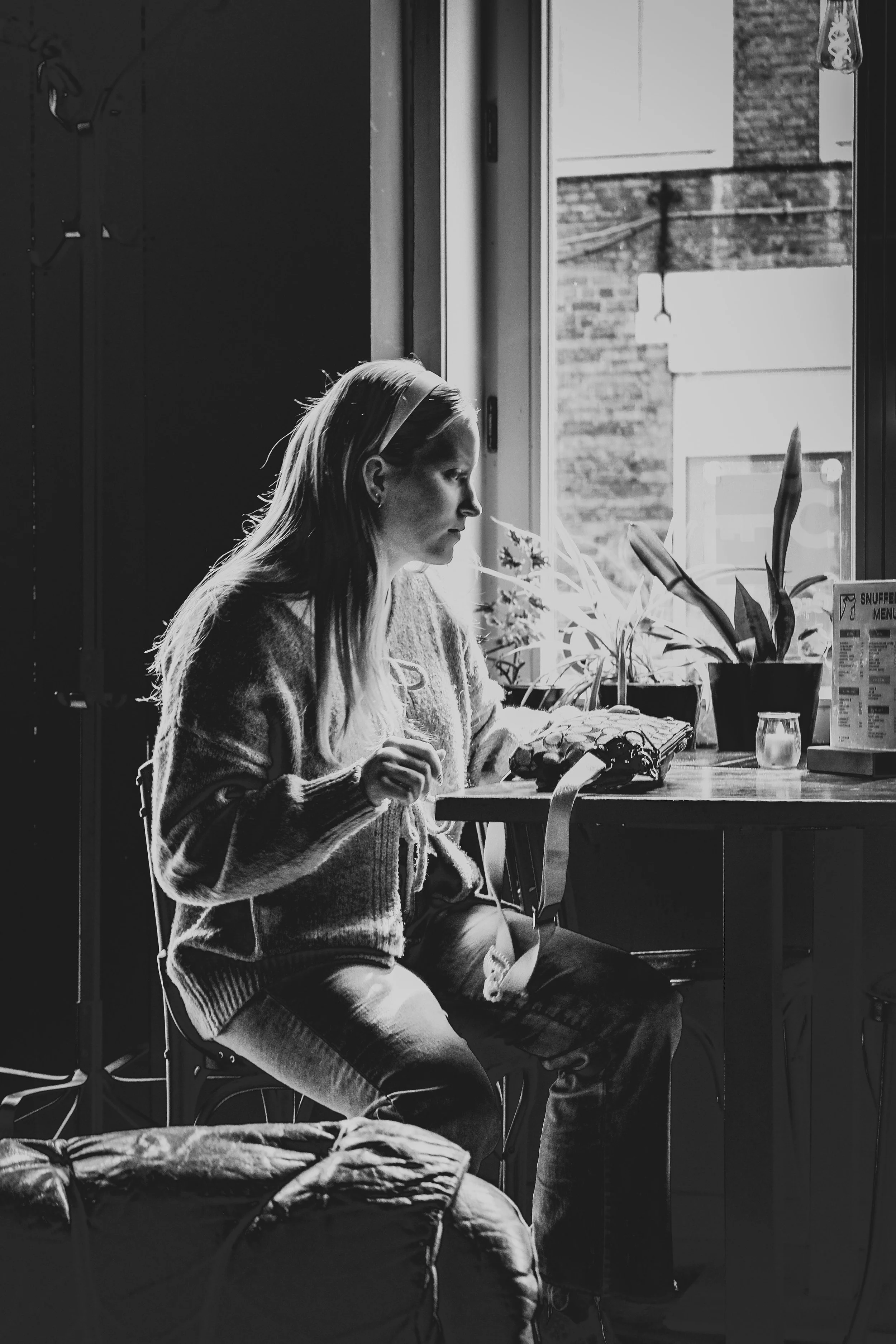 A woman sitting at a table near a window in a cafe, with plants and a small candle on the table, captured in black and white.