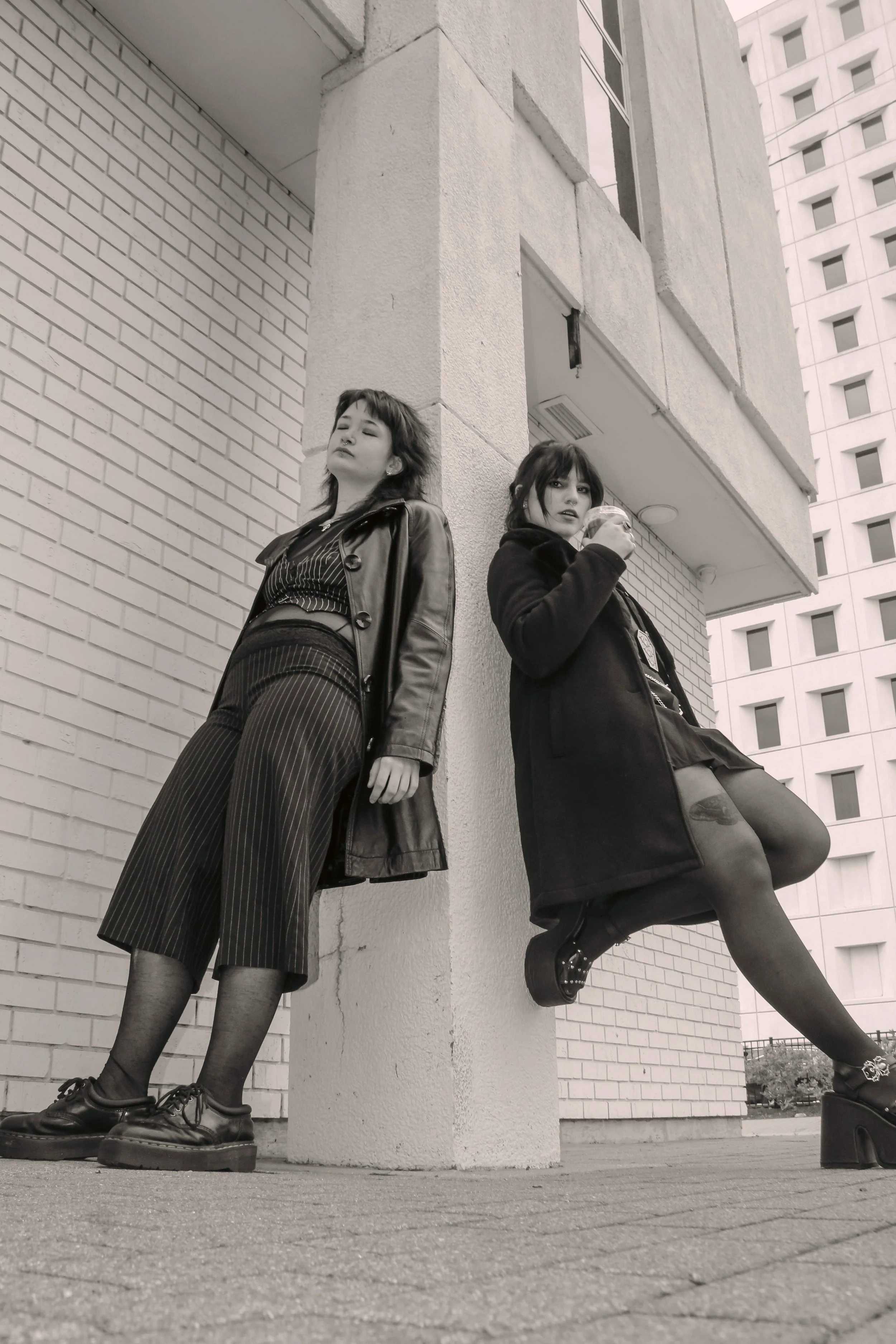 Black and white photo of two young women leaning against a corner of a building in an urban area. One woman is standing with her eyes closed, wearing a leather jacket and pinstripe pants, while the other woman is sitting on the ledge, drinking from a