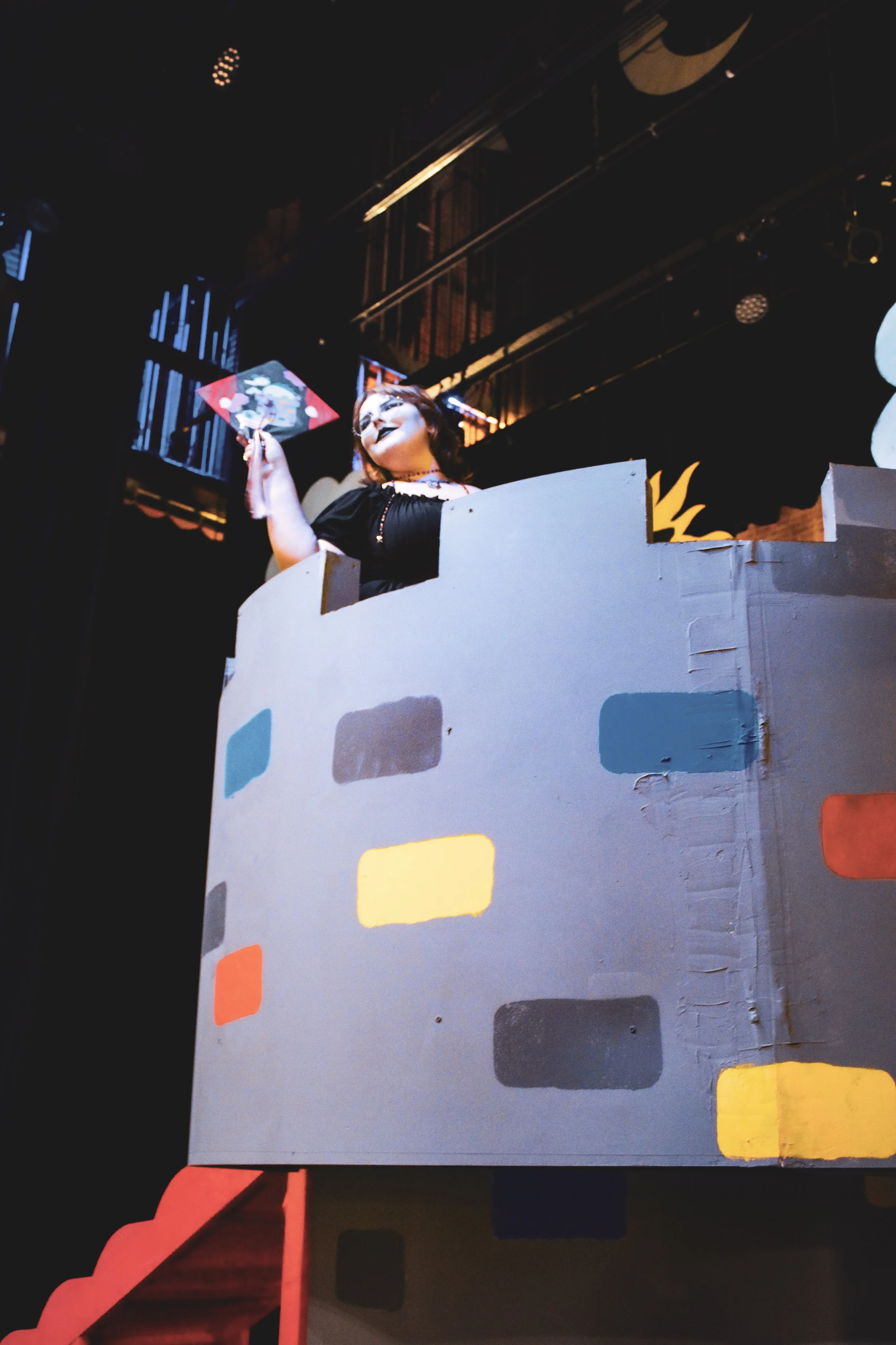 Person with dark makeup and dark hair holding a colorful art piece in a dimly lit room with colorful blocks on a structure