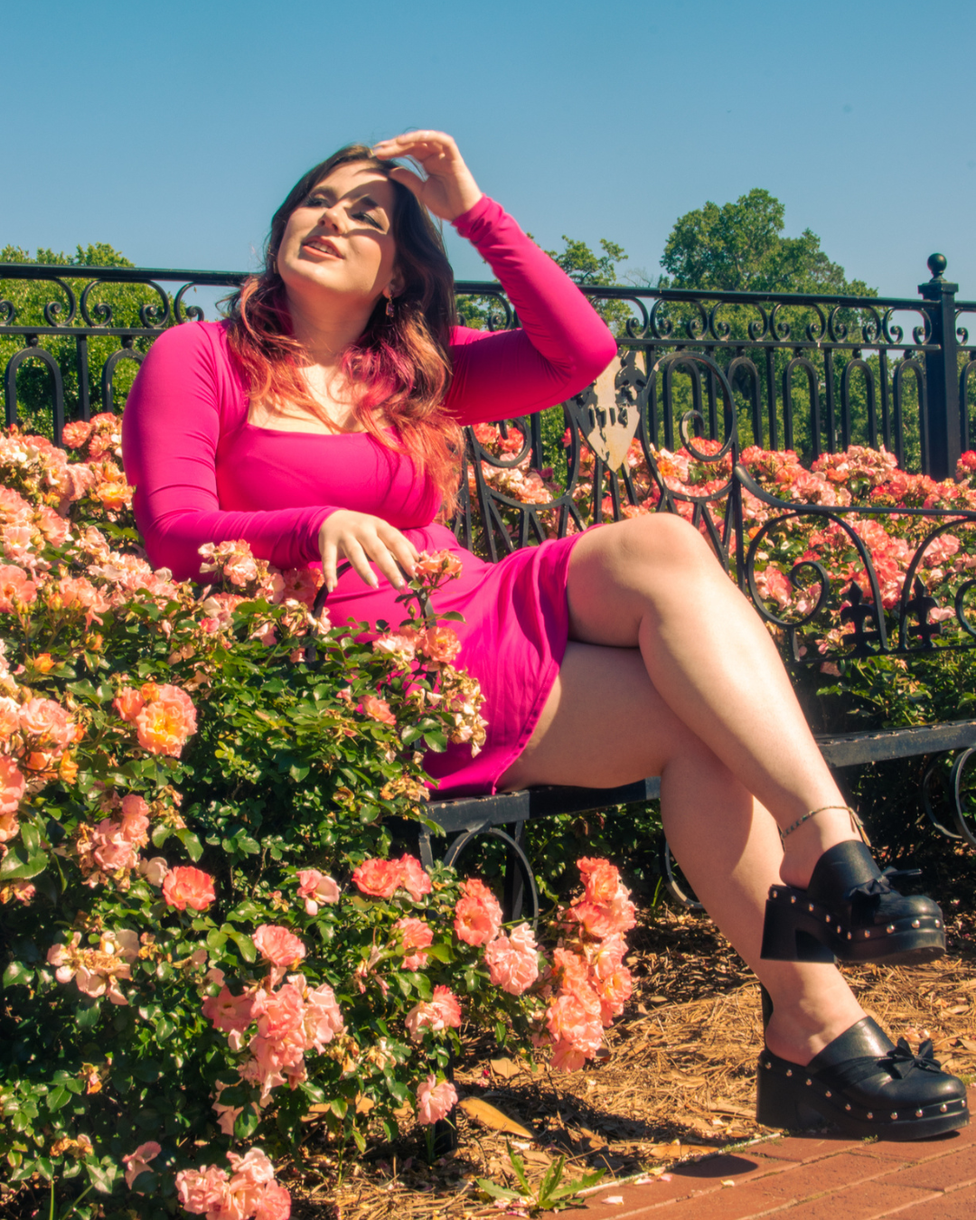Woman sitting on a park bench surrounded by pink flowers, wearing a pink dress and black platform shoes, shielding her eyes from the sun.