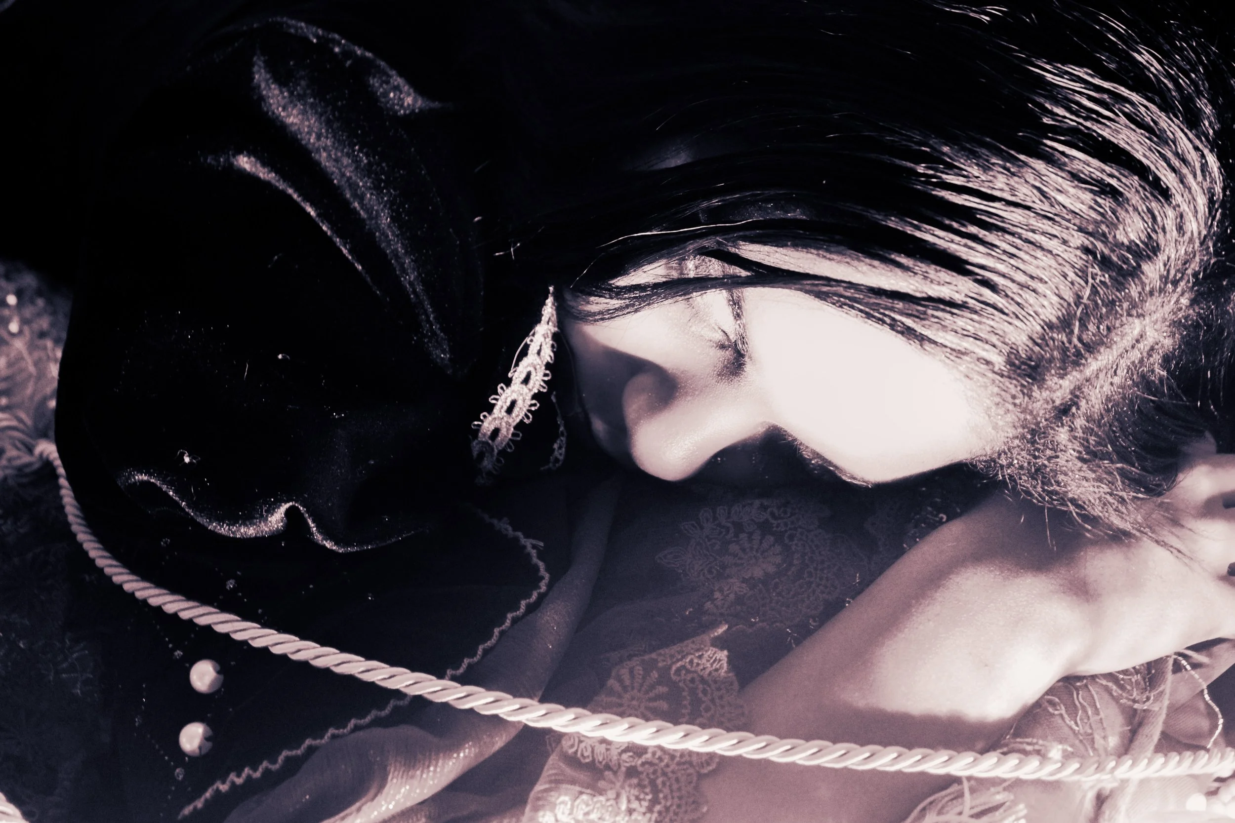A person with dark hair resting their face on their arm, dressed in dark, ornate clothing with lace and decorative trim.