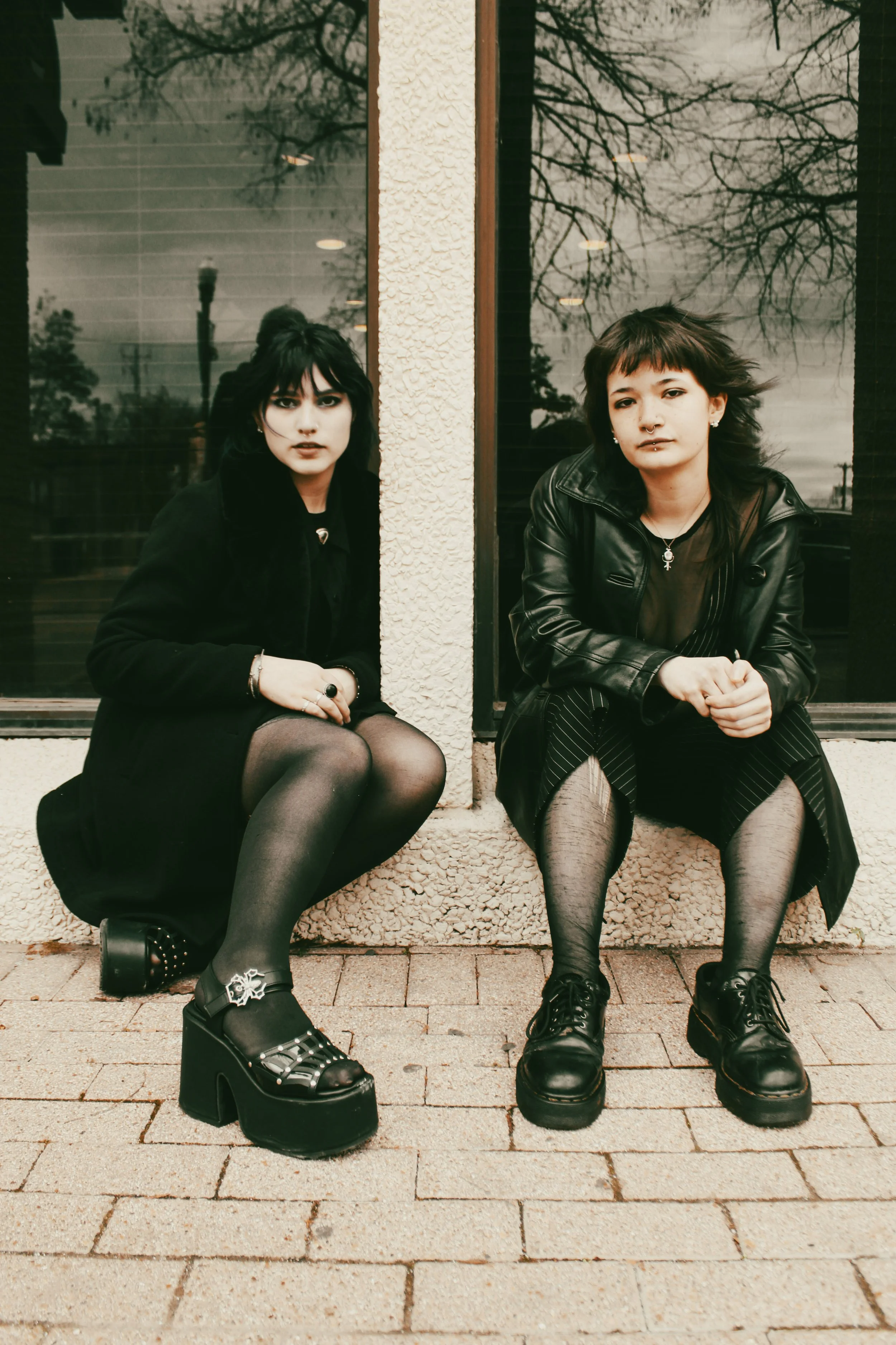 Two young women sitting on the sidewalk outside a building with large window reflections. They have dark hair, goth-style clothing, and platform shoes.