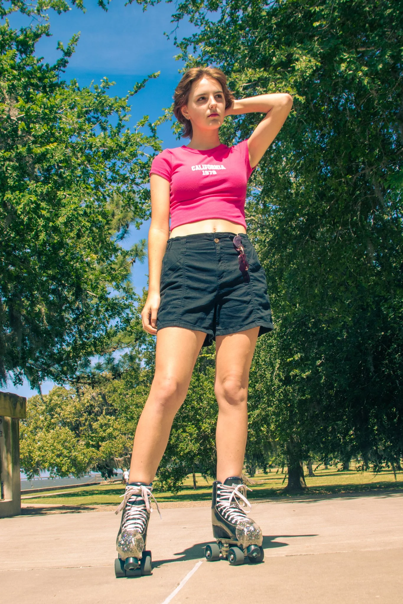 Young woman wearing a pink crop top and black shorts, roller skating outdoors on a sunny day, with trees and a blue sky in the background.