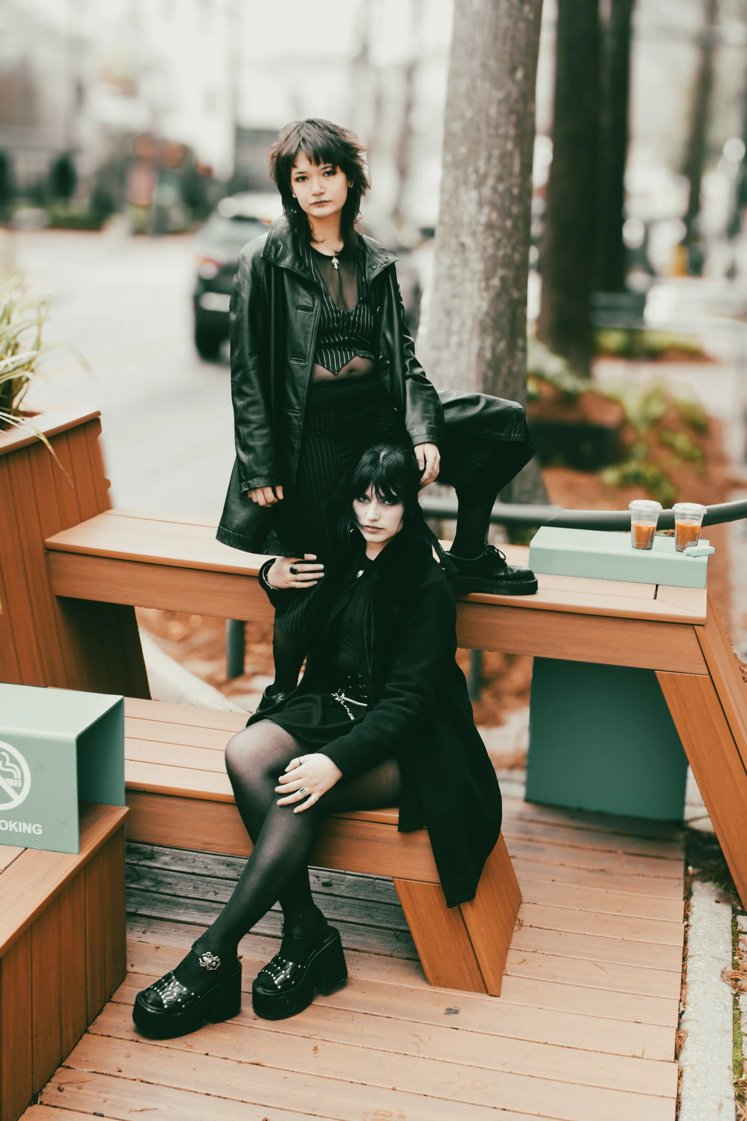 Two women dressed in black outfits sit and lean on a wooden park bench outdoors near trees and a sidewalk, with parked cars in the background.