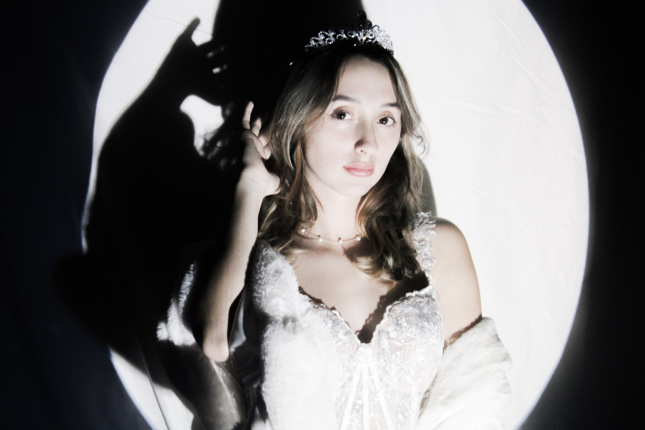A woman with wavy brown hair wearing a tiara and a lace dress, touching her earring, with her shadow cast on a circular light backdrop.