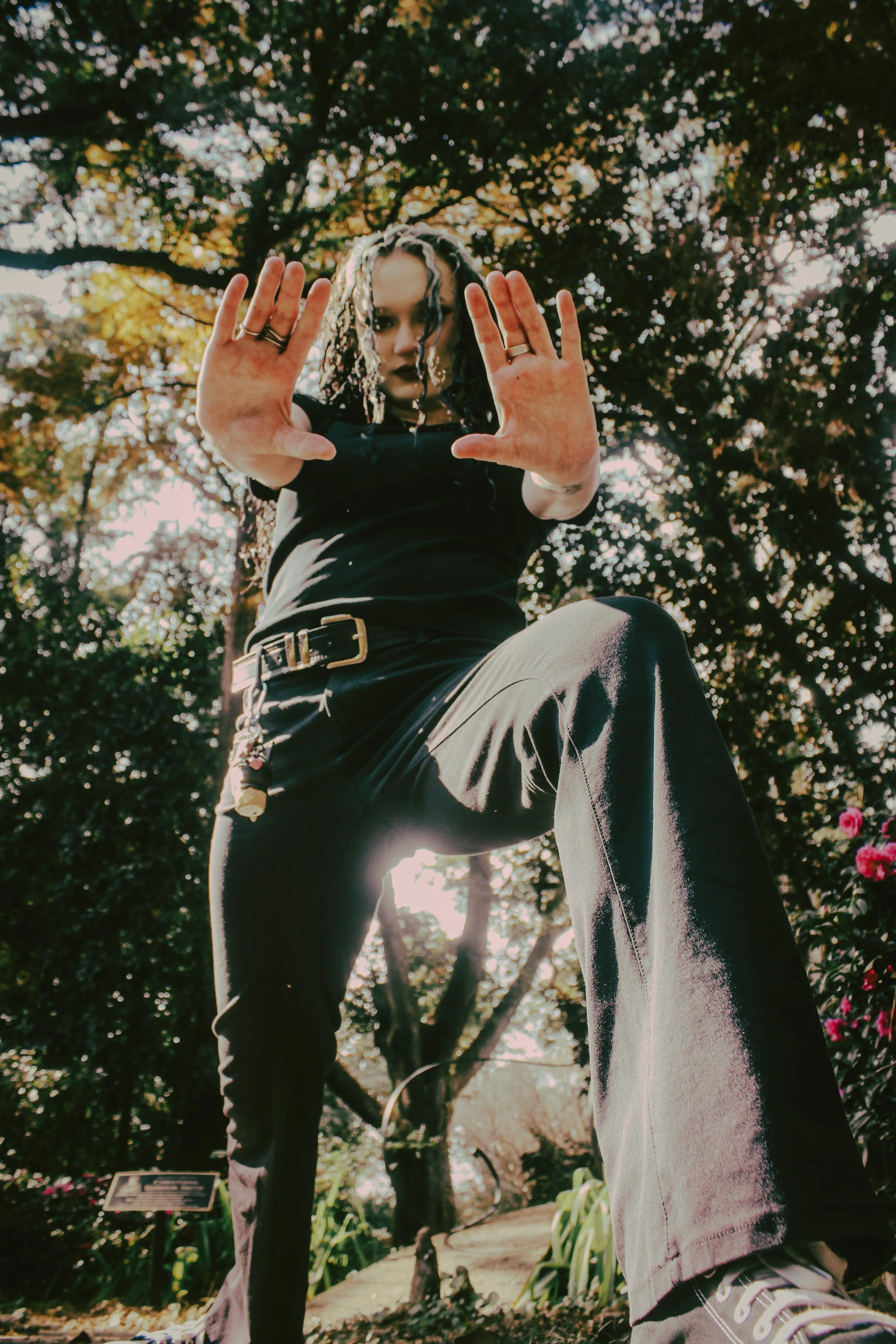 A person with dark hair, dressed in black, extending their hands towards the camera in a low-angle shot. They are outdoors among trees and flowers, with sunlight filtering through the leaves.