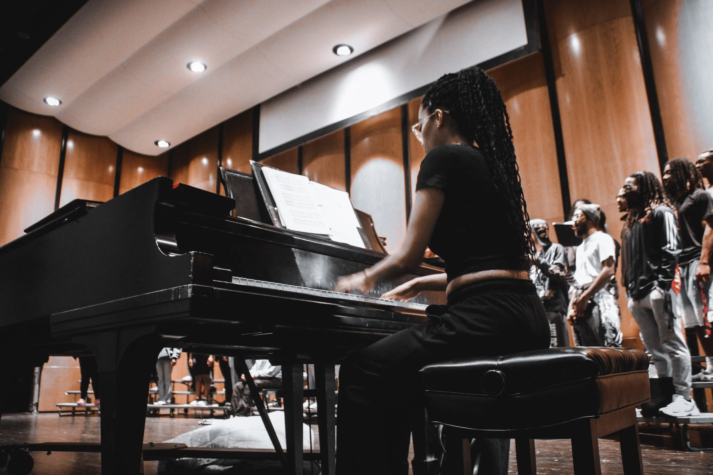 A woman playing a grand piano during a choir rehearsal or performance, with a choir singing in the background in a concert hall with wooden walls.