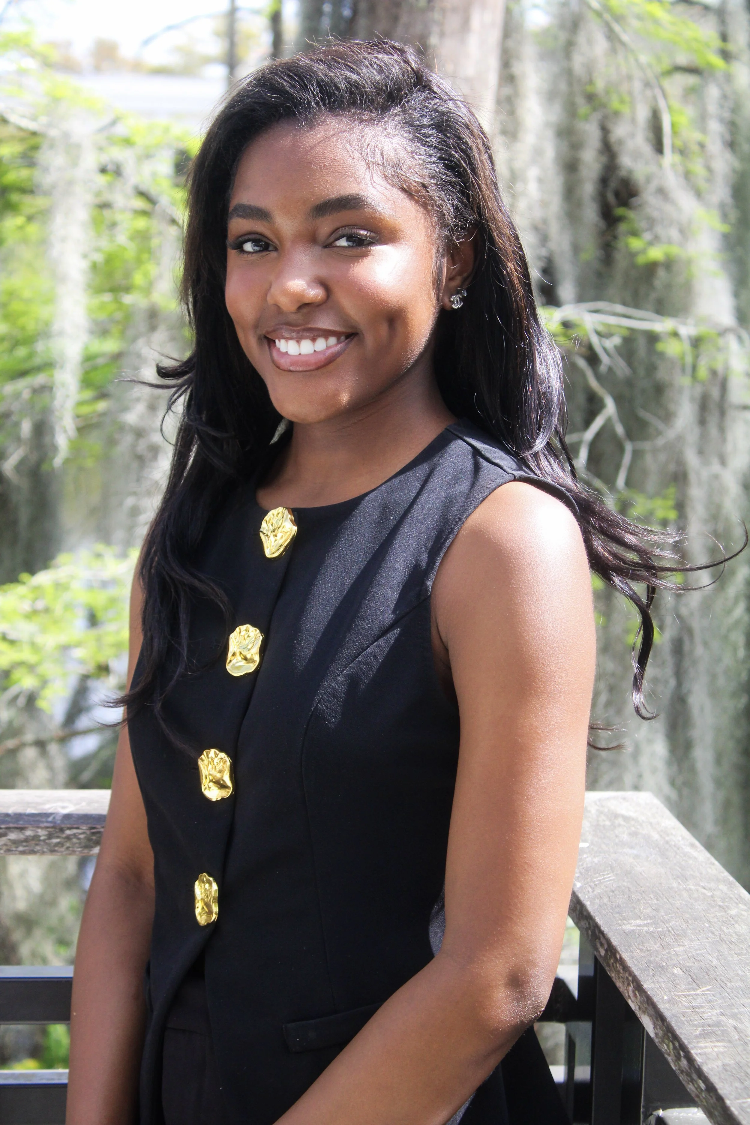 A woman with dark hair and earrings smiling outdoors in front of trees and a wooden railing.