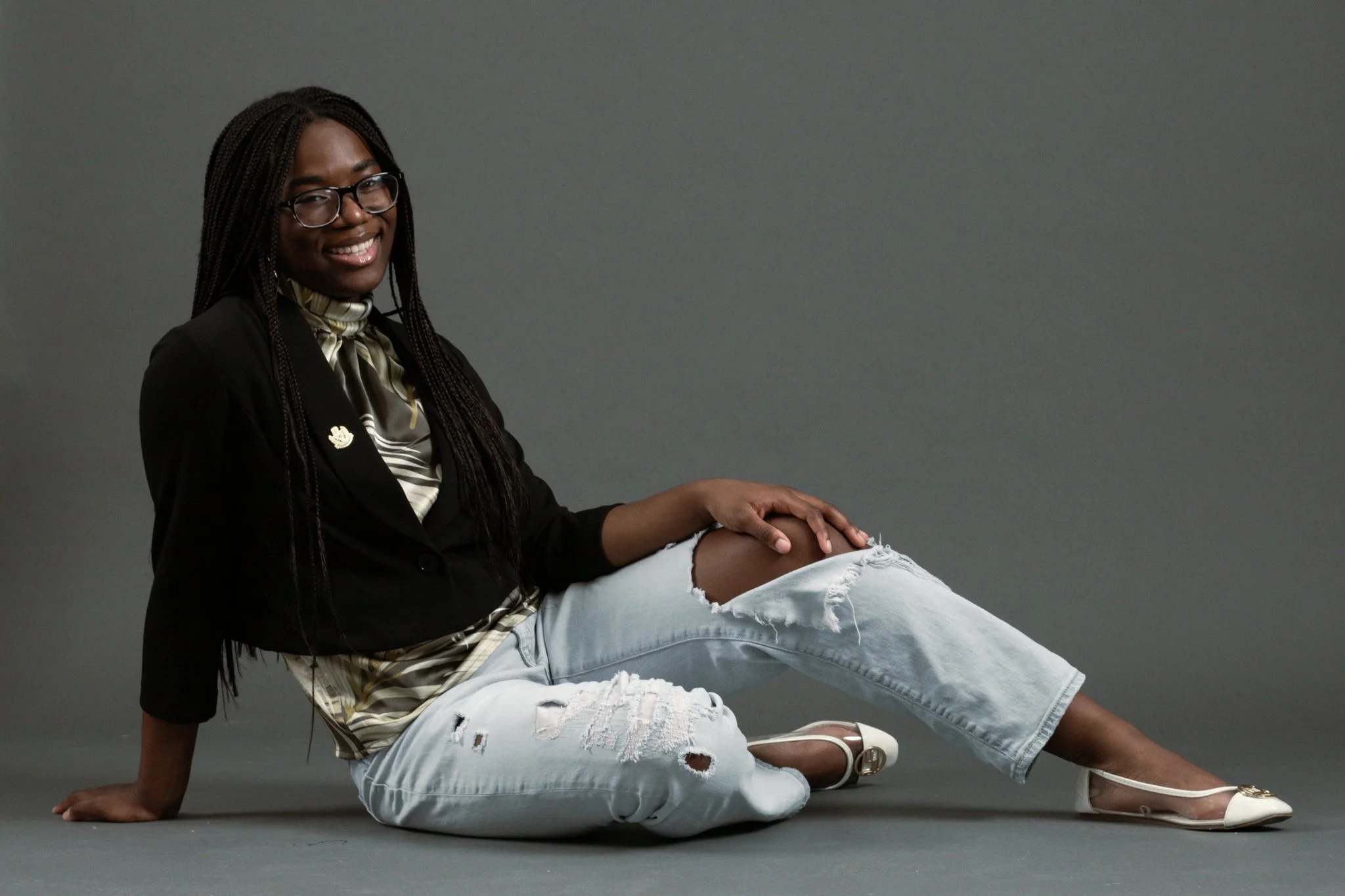 A smiling Black woman with glasses sitting on a gray floor against a gray background, wearing a black blazer, a patterned blouse, ripped light jeans, and white pointed-toe heels.