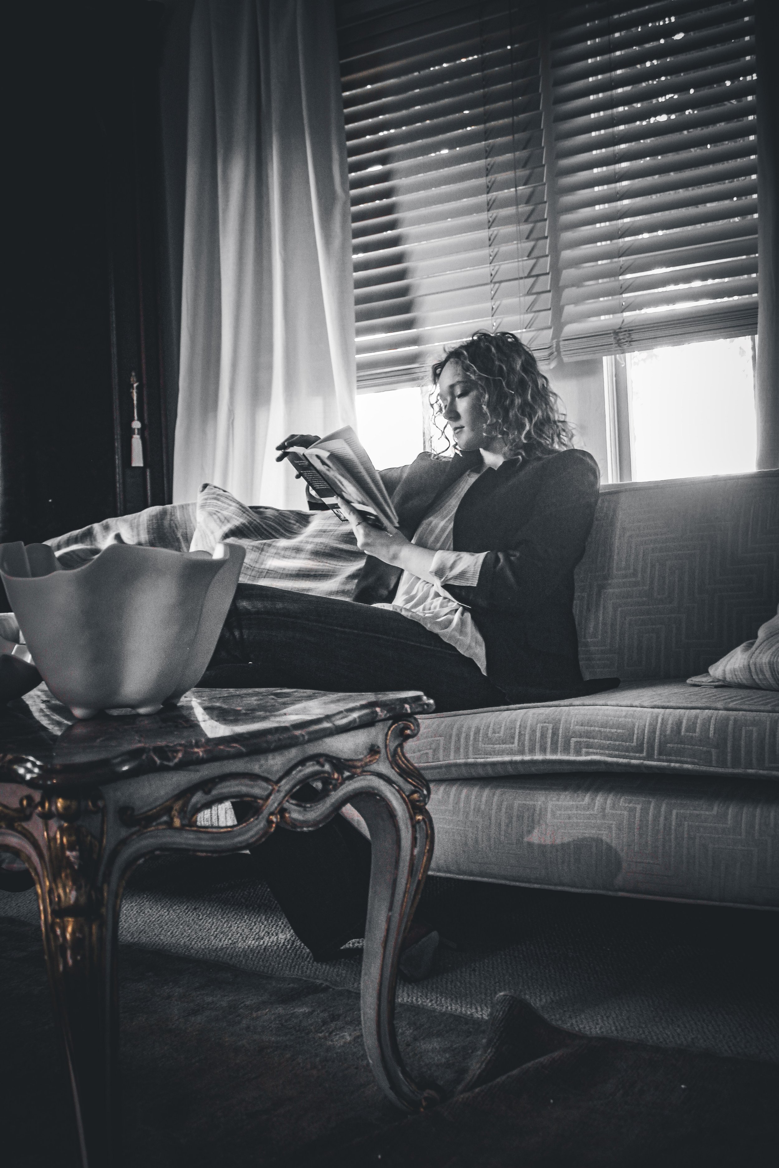 A woman with curly hair reading a book or magazine while sitting on a couch in a living room, with window blinds and curtains behind her, and a decorative table in front.
