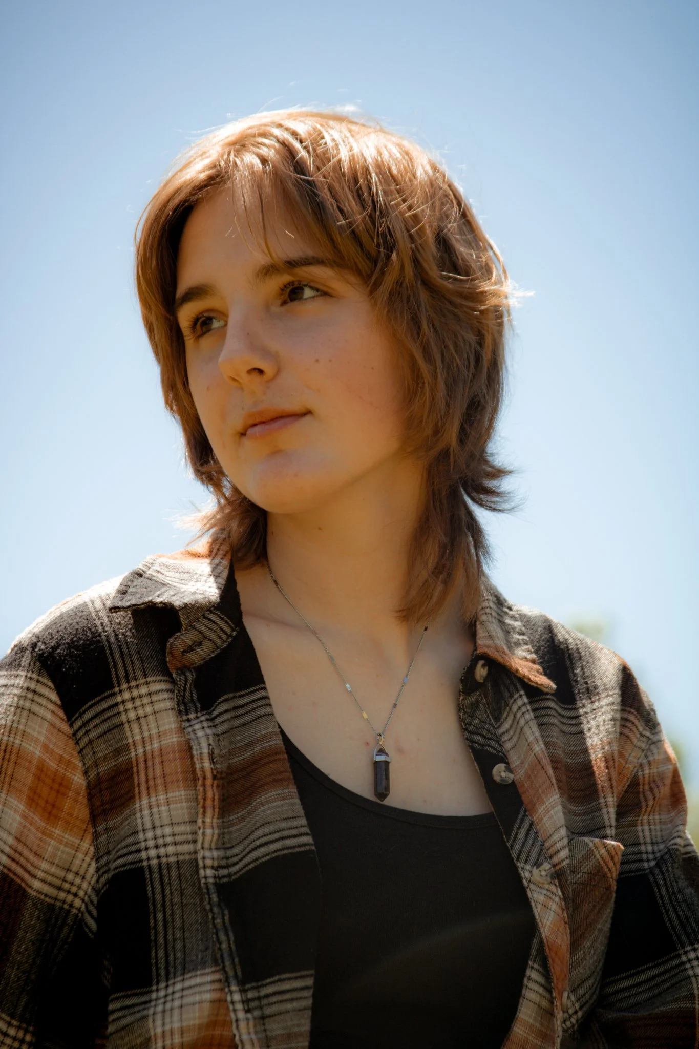 A young woman with short, wavy, reddish-brown hair is outdoors under a clear blue sky, wearing a plaid shirt over a black top and a pendant necklace.