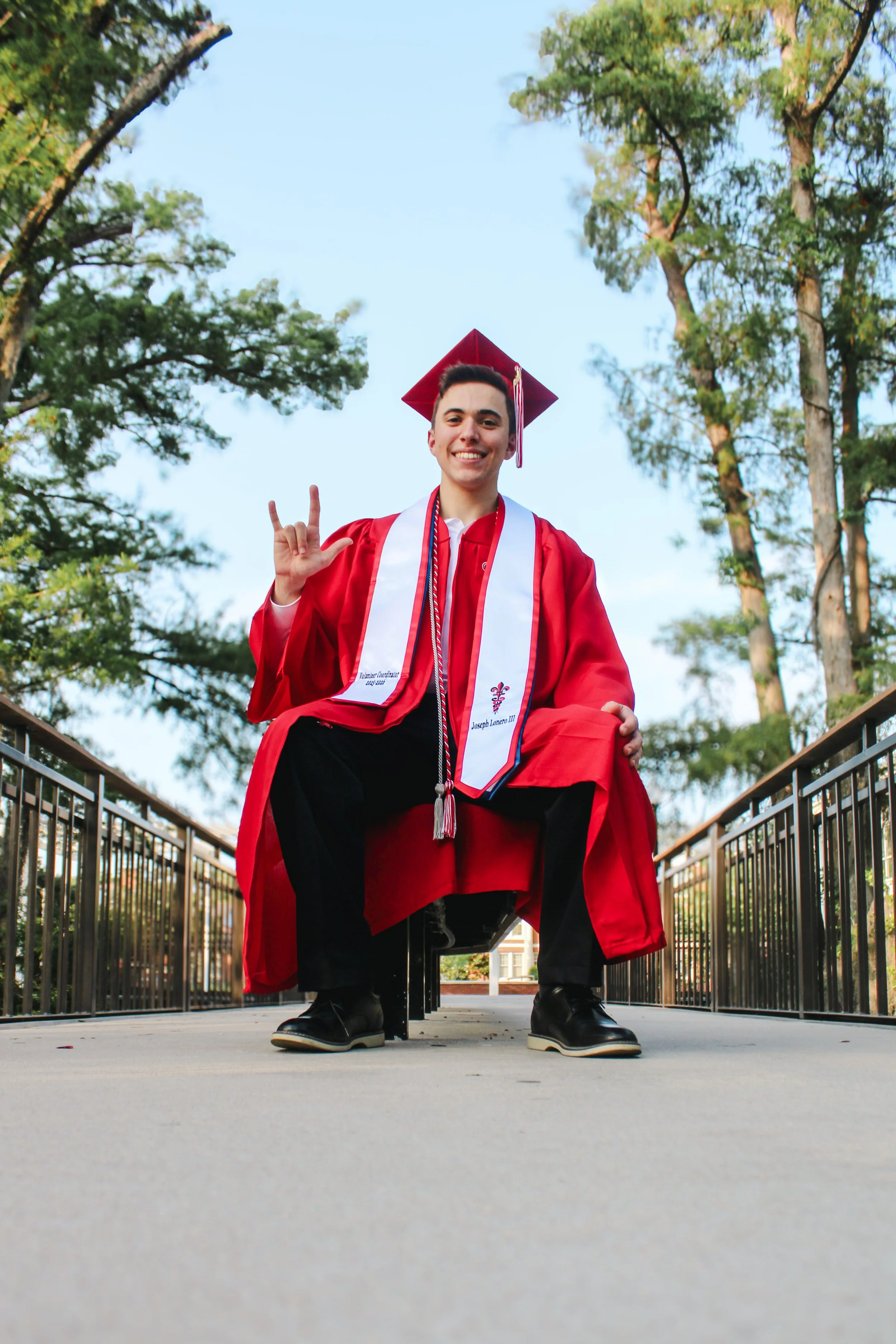 Young man in a red graduation gown and cap, sitting on a ledge outdoors, showing a 'rock on' hand gesture, smiling, with tall trees and a blue sky in the background.