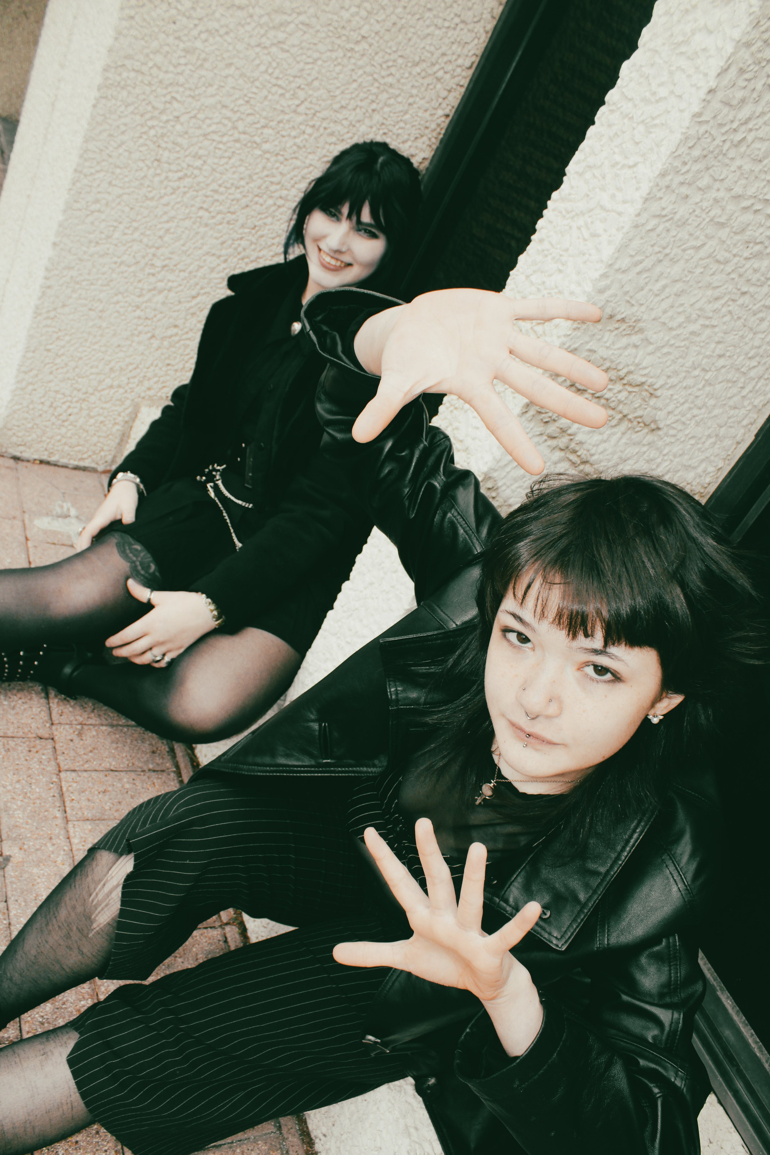 Two young women sitting against a wall, reaching towards the camera, smiling, dressed in black leather jackets, skirts, and tights, with dark hair and piercings.