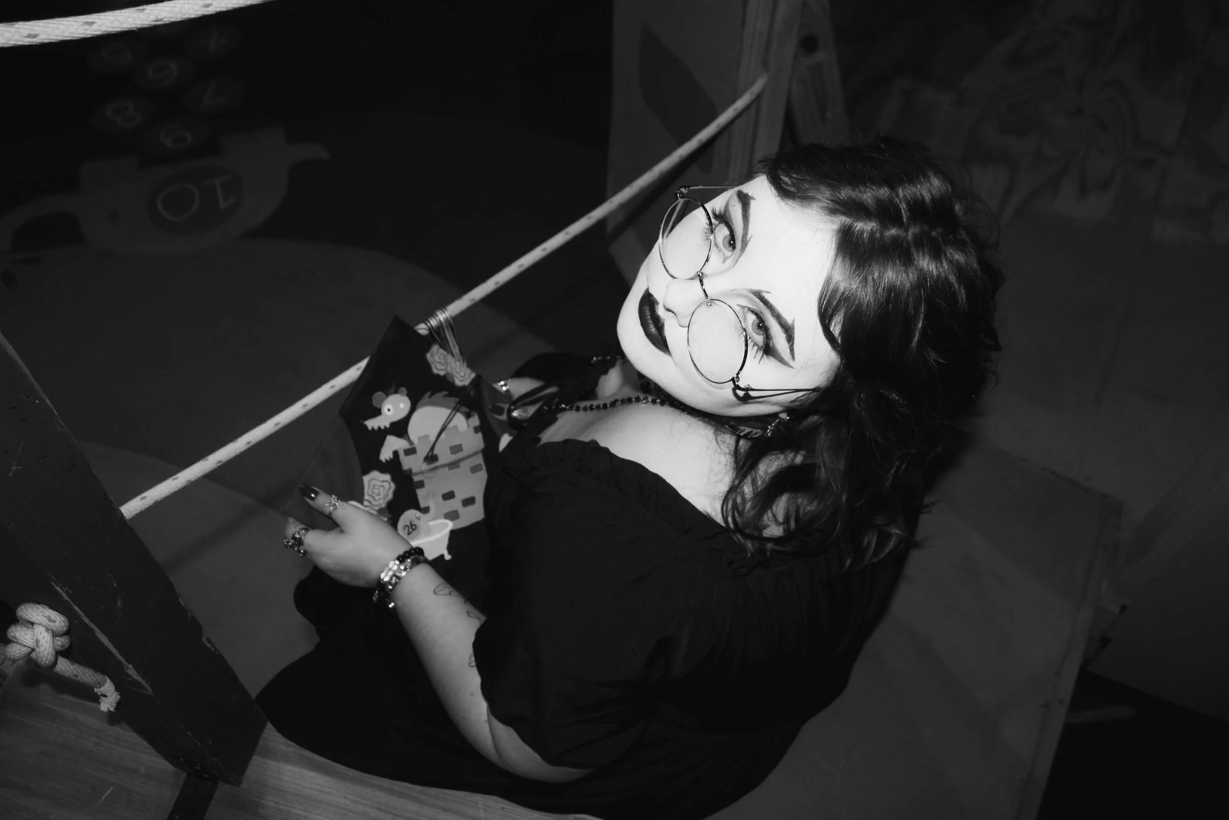 A woman with dark wavy hair and glasses, dressed in dark clothing, holding a small bag with a Halloween design, sitting against a wall with a Halloween-themed mural or decoration, in a black-and-white photograph.
