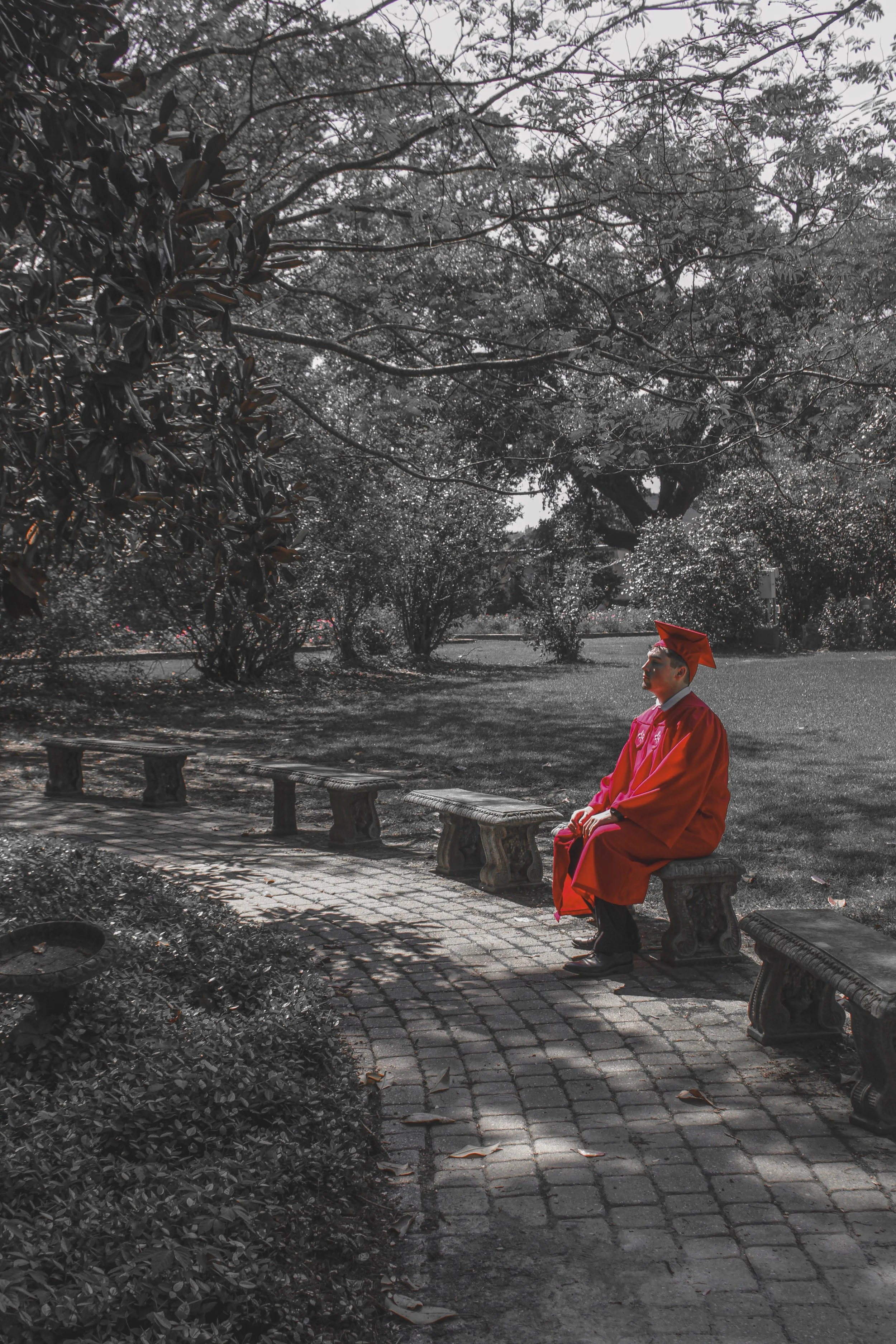 A person dressed in a bright red graduation gown and cap is sitting on a park bench, looking to the side. The background is in black and white, featuring large trees, bushes, and a cobblestone pathway.