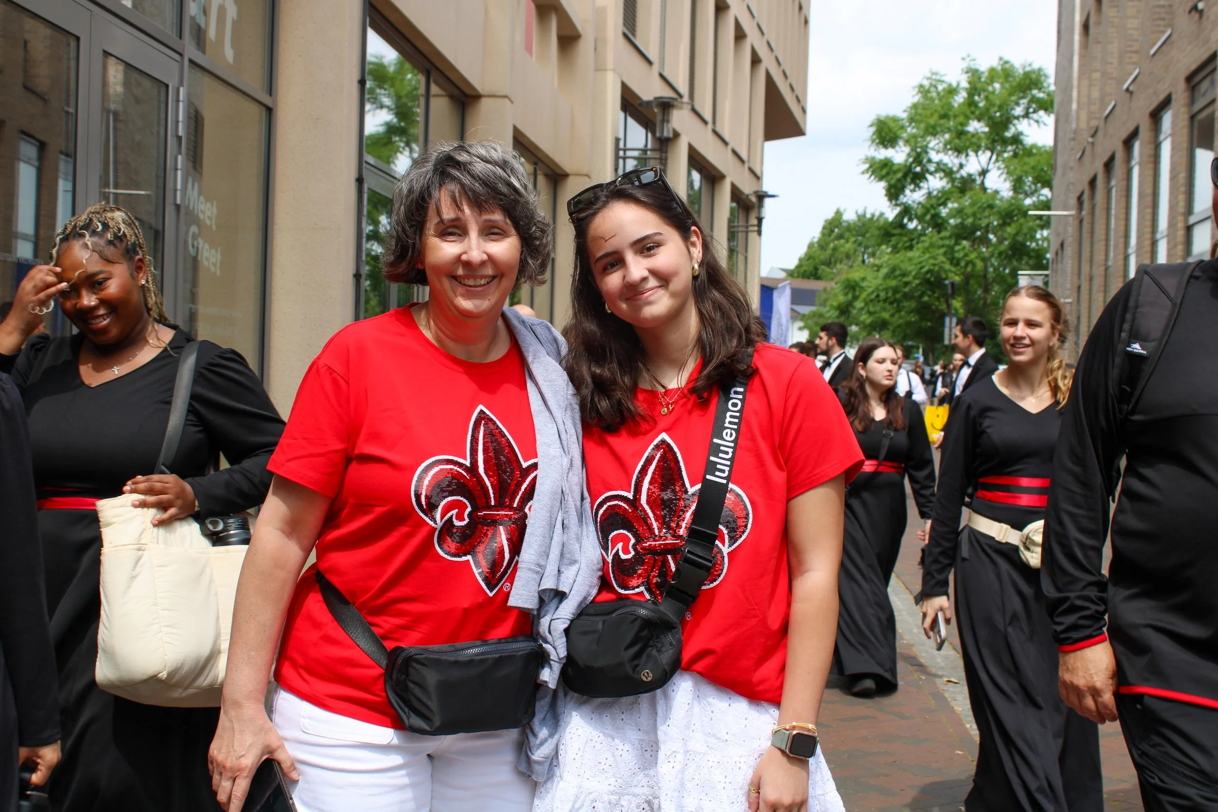 Two women with gray and dark brown hair standing together outside in a festive setting, both wearing red shirts with a black and white fleur-de-lis logo, smiling at the camera. Other people are visible in the background, some dressed in black and red