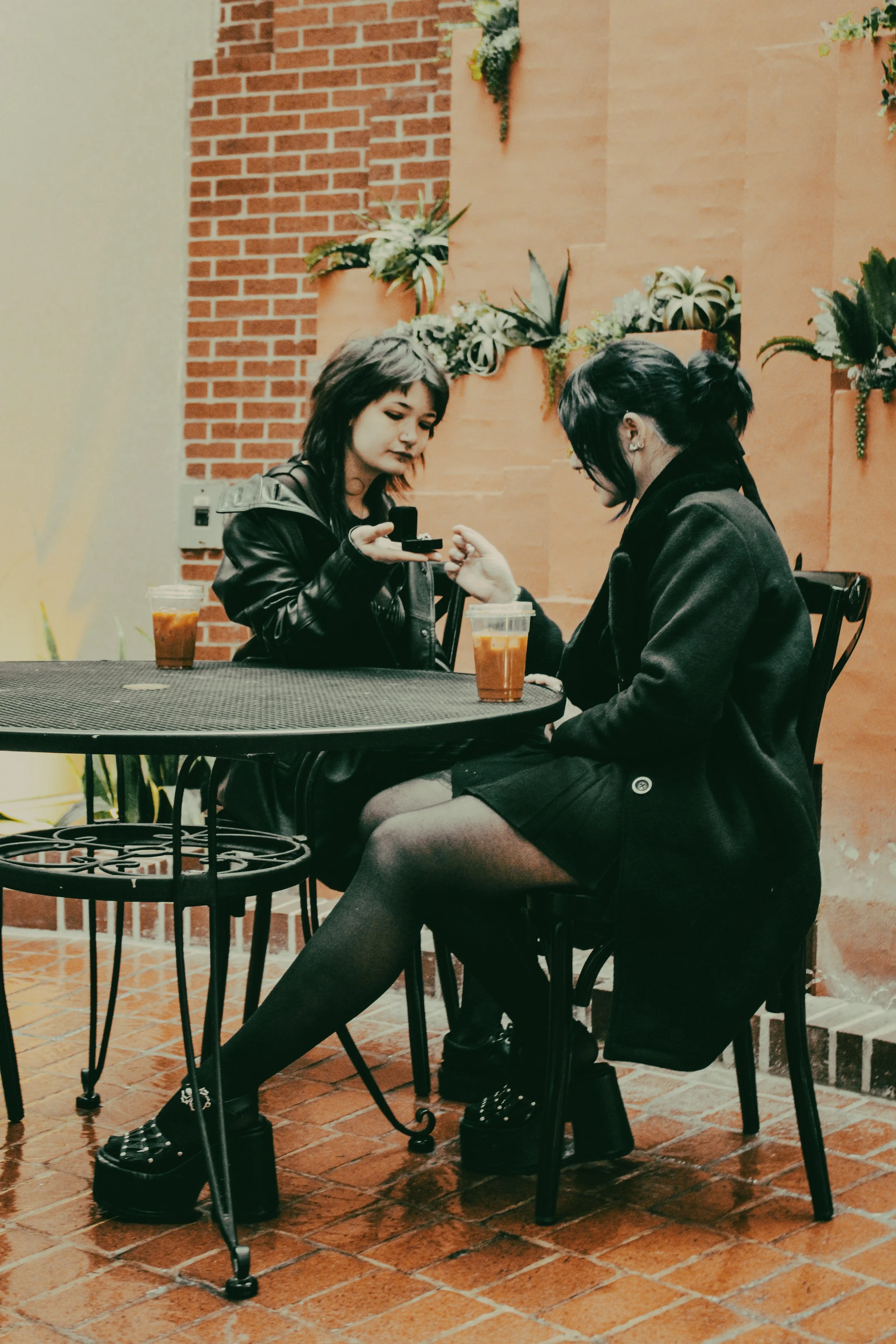 Two young women sit at a black metal table with drinks in plastic containers, inside a cafe with orange tiled walls and indoor plants. One woman shows something on her phone to the other.