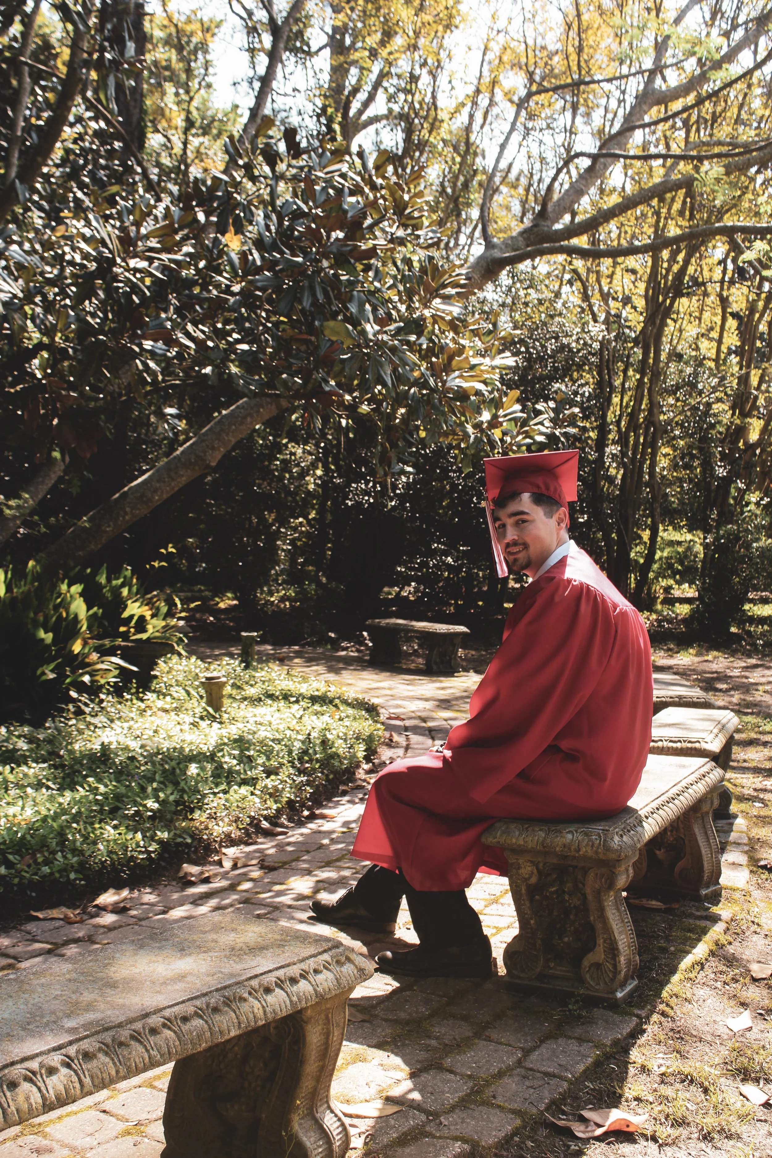 A young man in a red graduation gown and cap sitting on a stone bench in a park surrounded by trees with autumn leaves.