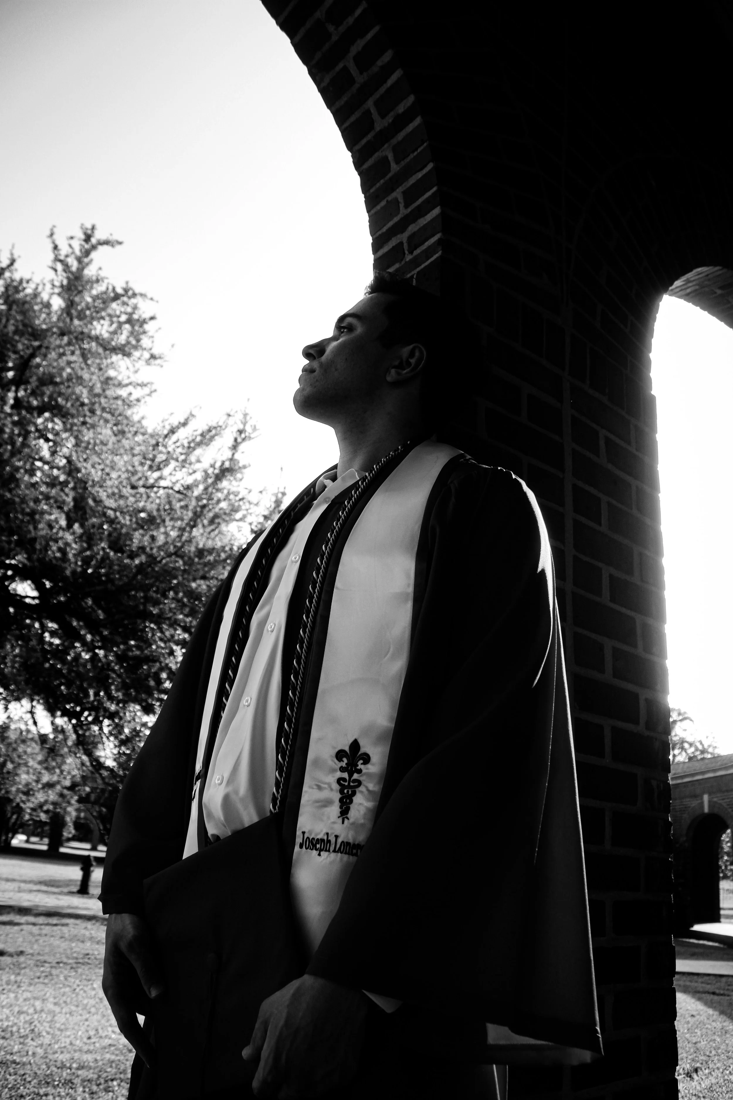 A young man in a graduation gown and stole, standing outdoors near a brick archway. The photo captures his profile as he looks upward, with sunlight backlighting him and trees in the background.