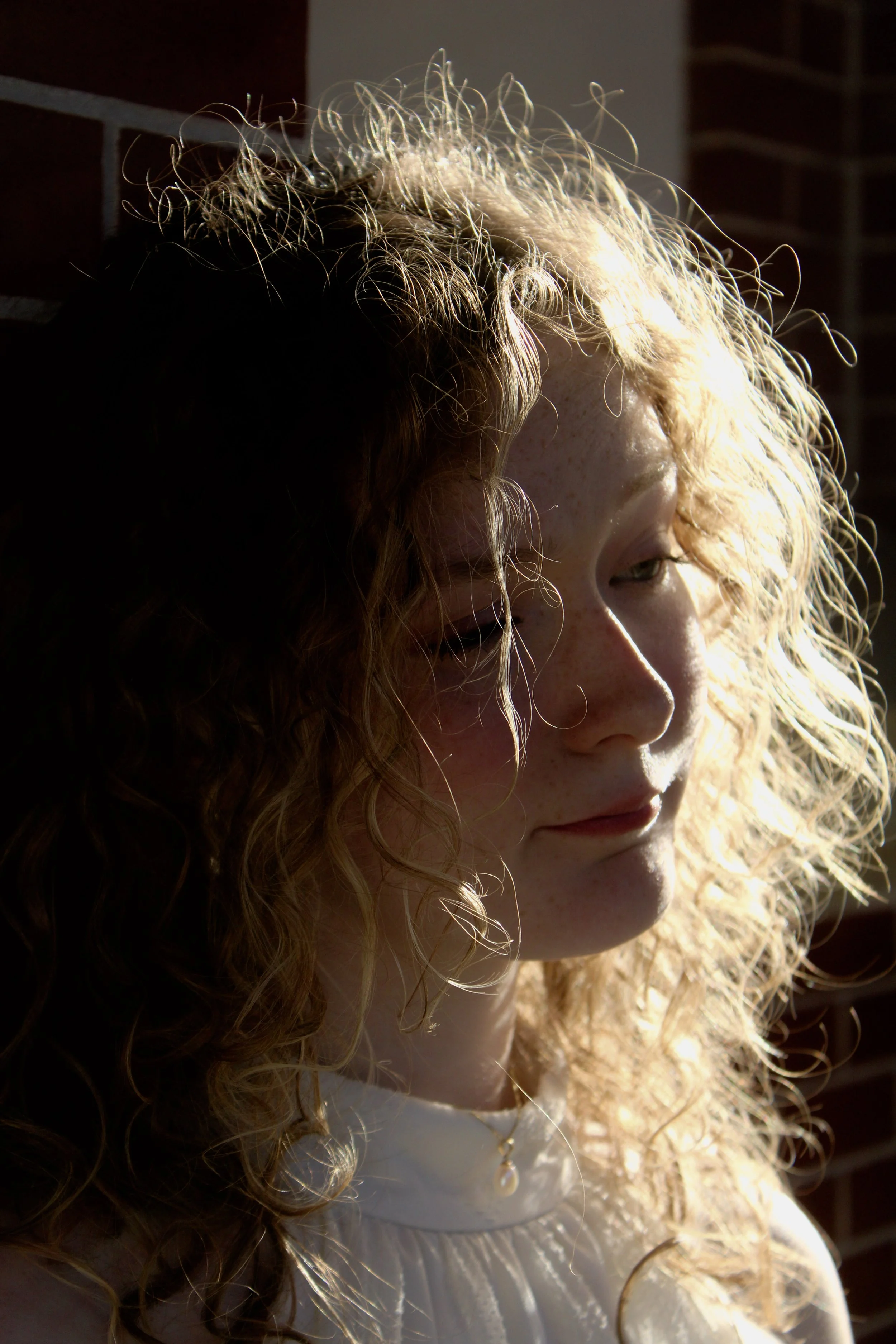A woman with curly blonde hair and light skin, wearing a white top, standing in sunlight with a dark background and brick wall.