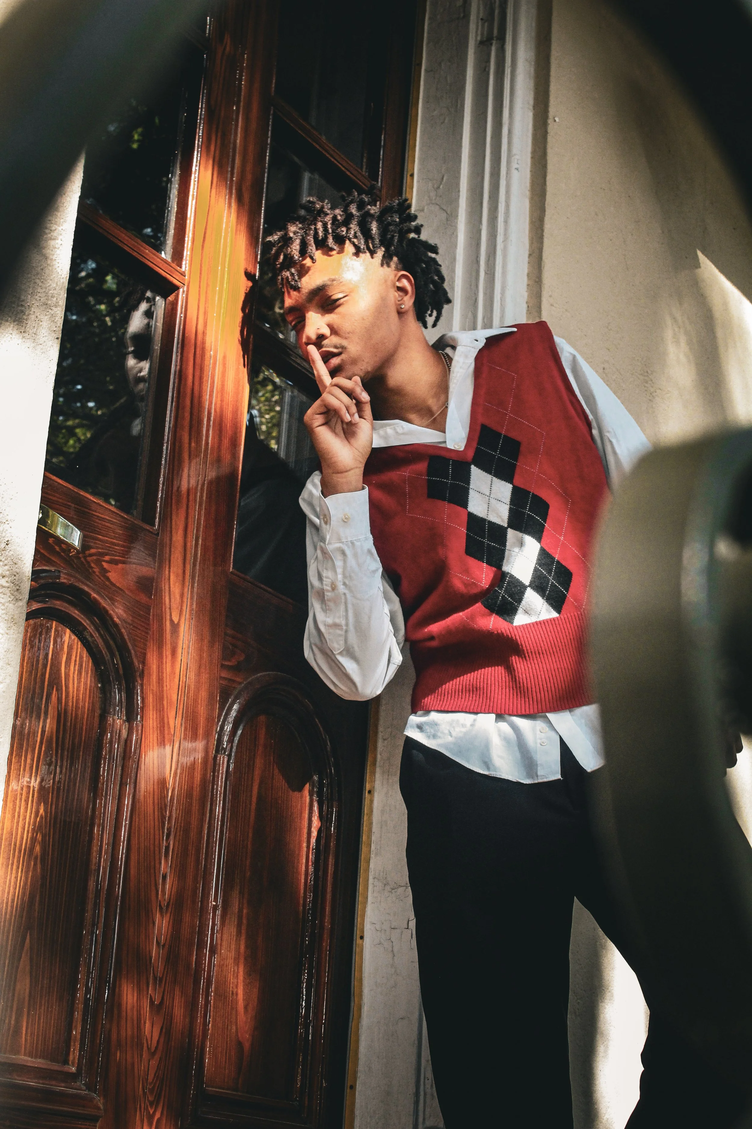 A young man with styled hair and earrings making a 'shh' gesture while leaning against a wooden door, dressed in a white shirt, red vest with black and white pattern, and black pants.