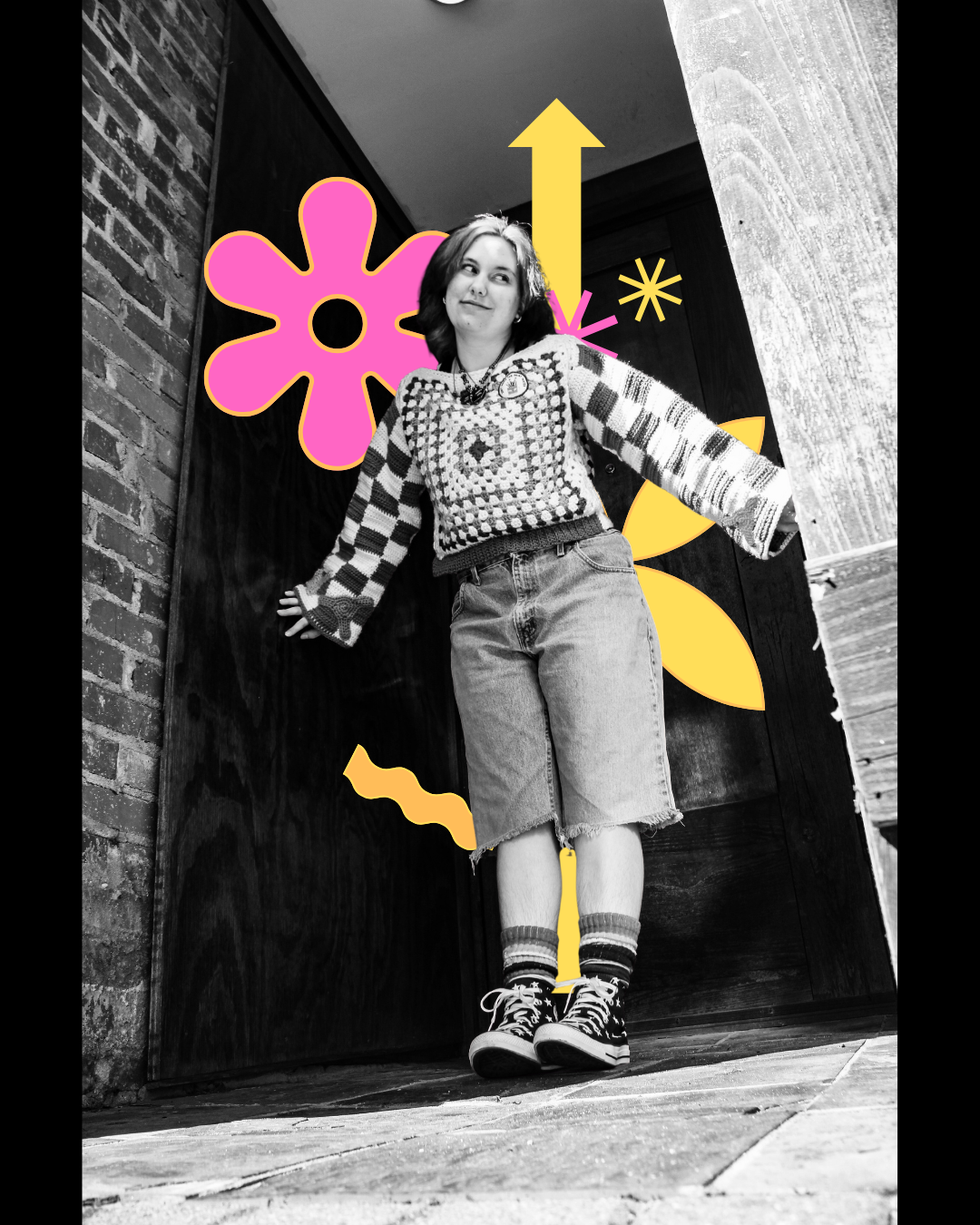Black and white photo of a young woman standing against a wall decorated with colorful, abstract graphic elements like a pink flower, yellow star, arrow, and other shapes.