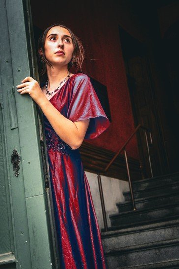Young woman in a red and purple dress standing near a doorway, looking outside, indoors with stairs behind her.