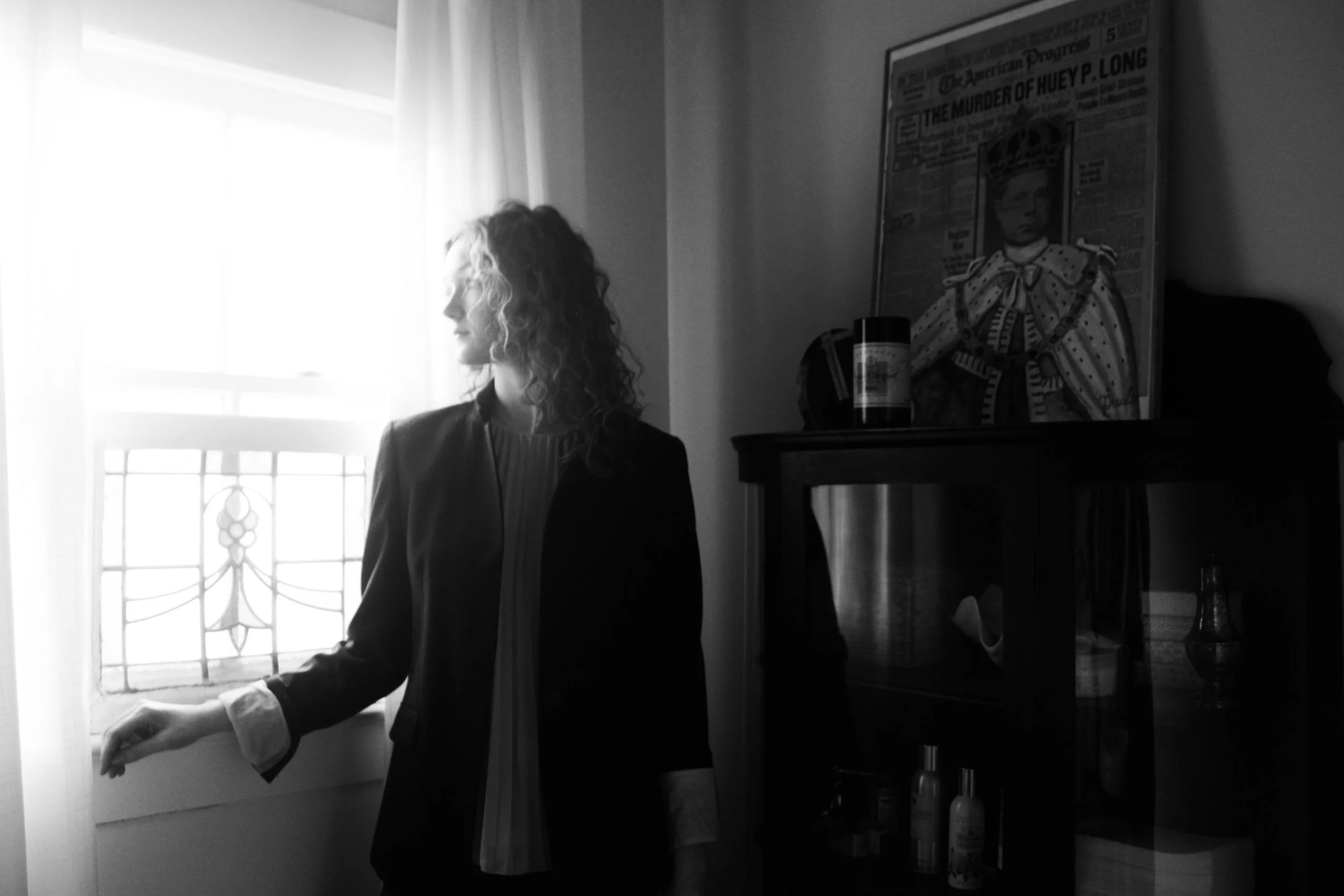 A woman with curly hair wearing glasses and a dark jacket, standing near a window with sheer curtains. Behind her, a framed artwork of Frida Kahlo is displayed on a dark wall.