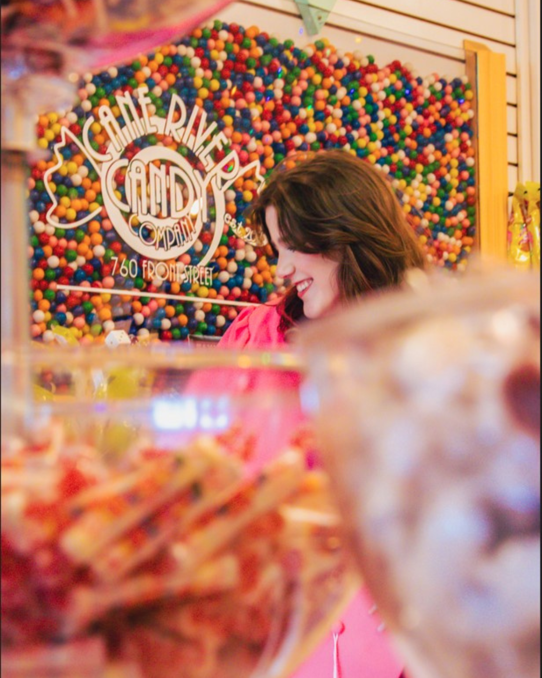 A woman smiling at a candy store, with a background of multicolored gumballs, and a sign that reads "ACME RIVER CANDY COMPANY, 760 FRONT STREET."