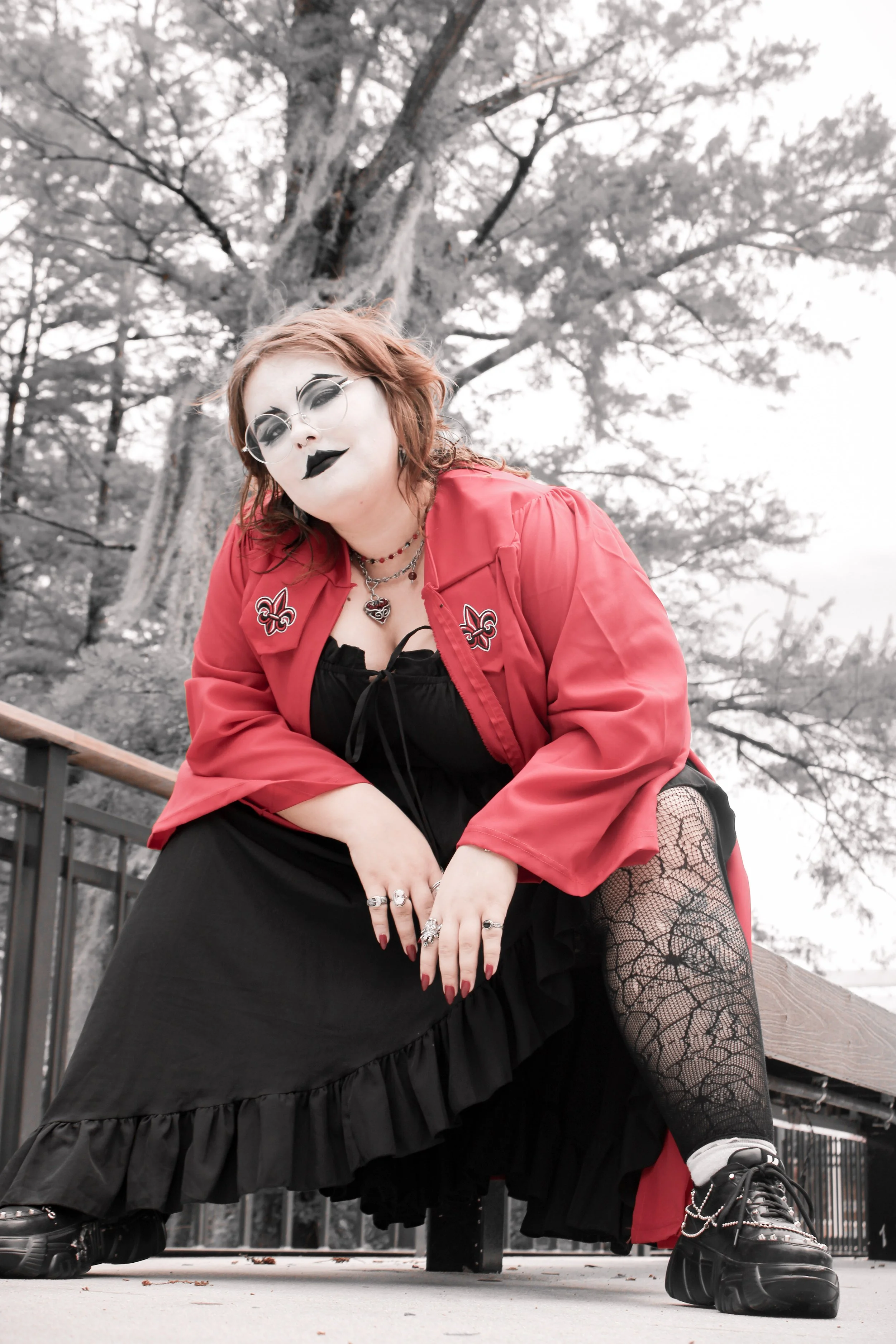 A woman with red hair and tattoos, dressed in gothic style, poses outdoors near a tree. She wears sunglasses, black lipstick, a red jacket with patches, layered necklaces, fishnet tights, black platform shoes, and dark makeup.