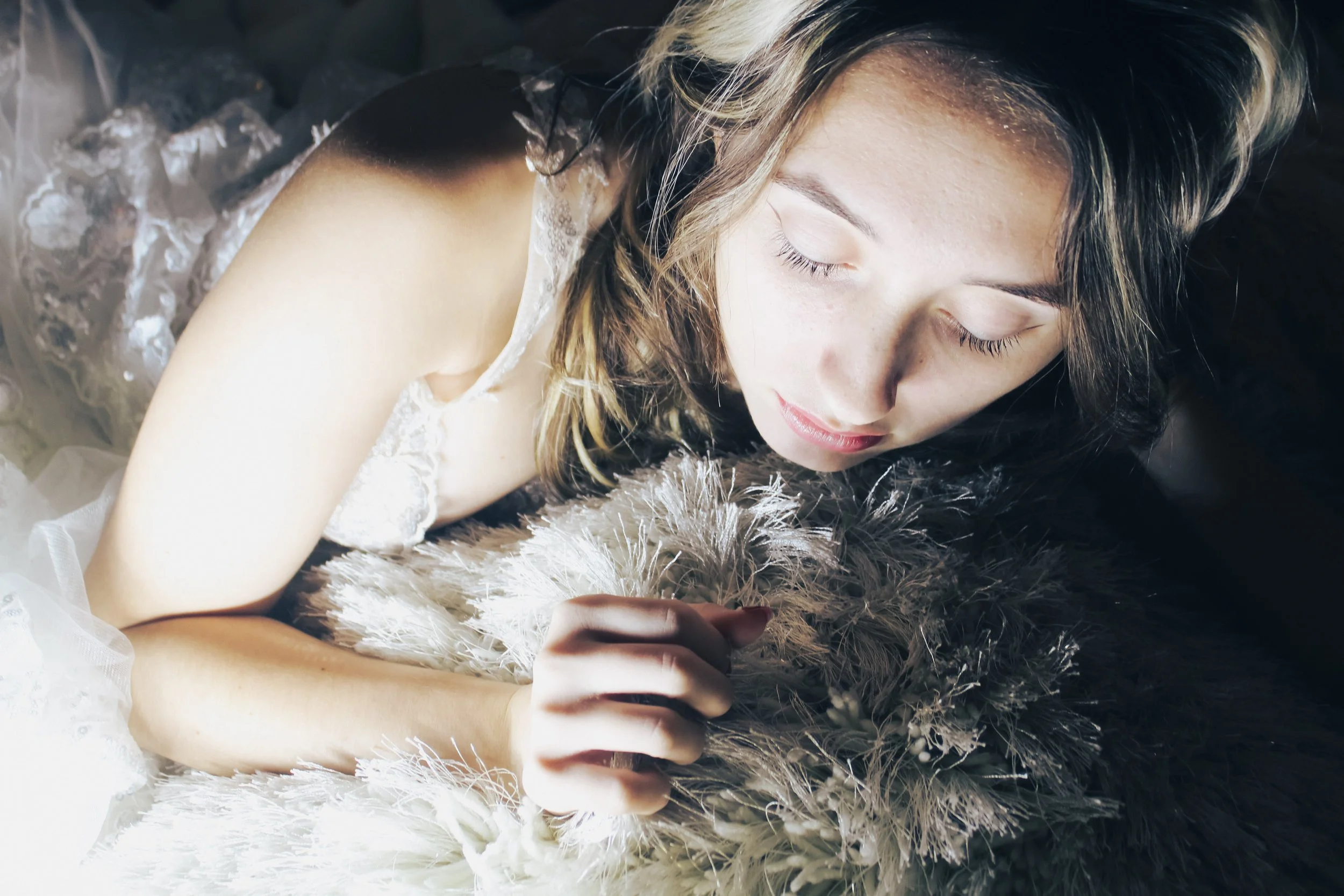 A young woman with closed eyes lying on a fluffy feathered surface, wearing a white lace dress.