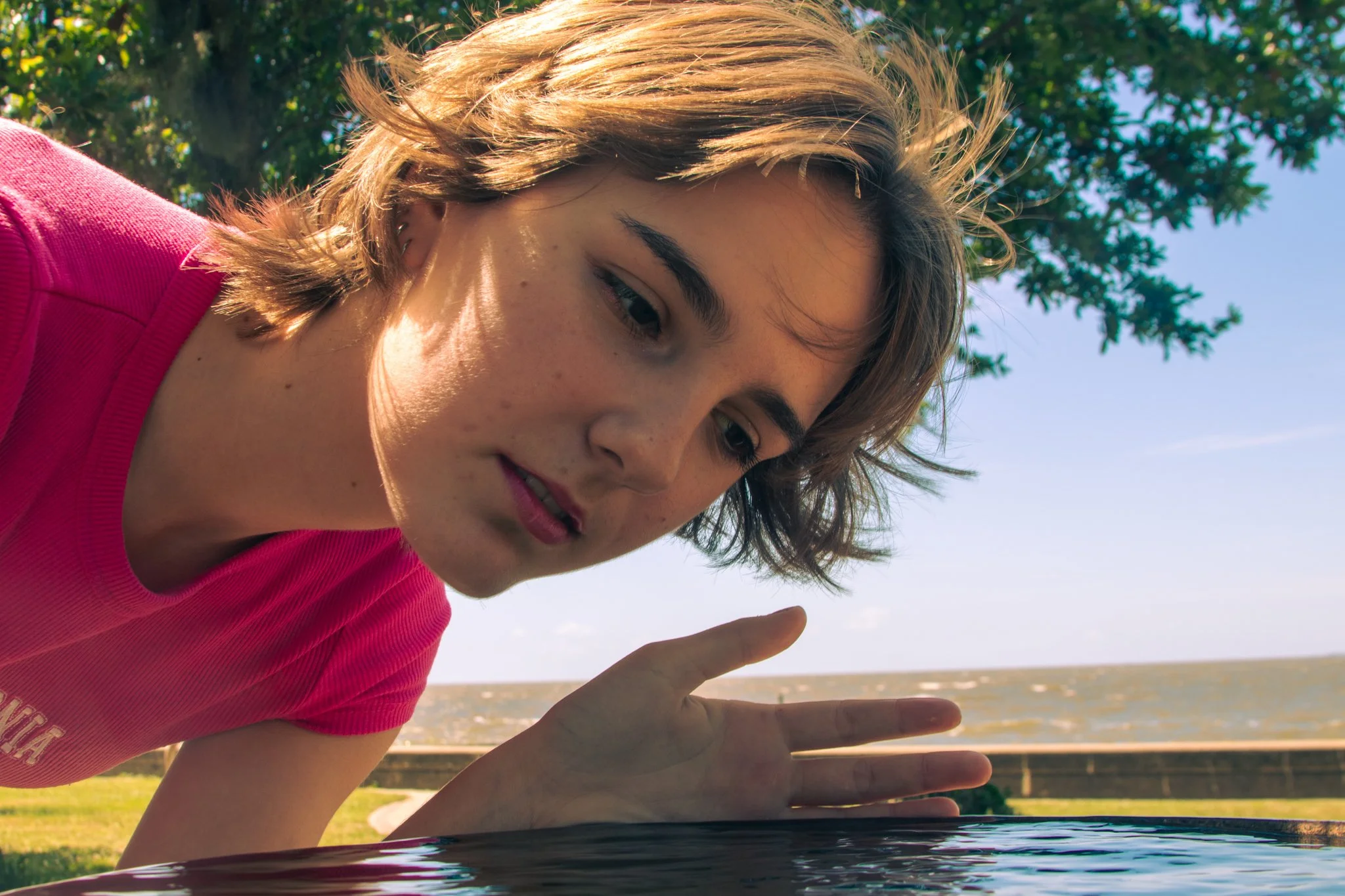 A young woman with short, wavy hair wearing a pink shirt leaning over water outdoors, with trees and sky in the background.